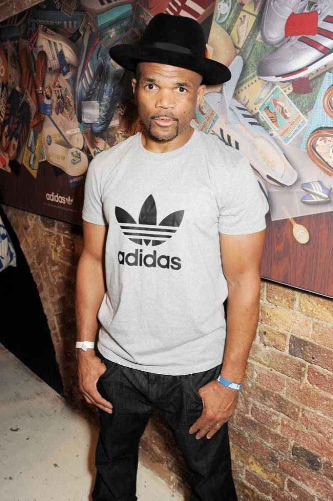 說唱歌手 桌布 壁紙 — 一個戴黑色軟呢帽、身穿灰色adidas T恤的男人自信地站在貼滿運動鞋海報的磚牆前。