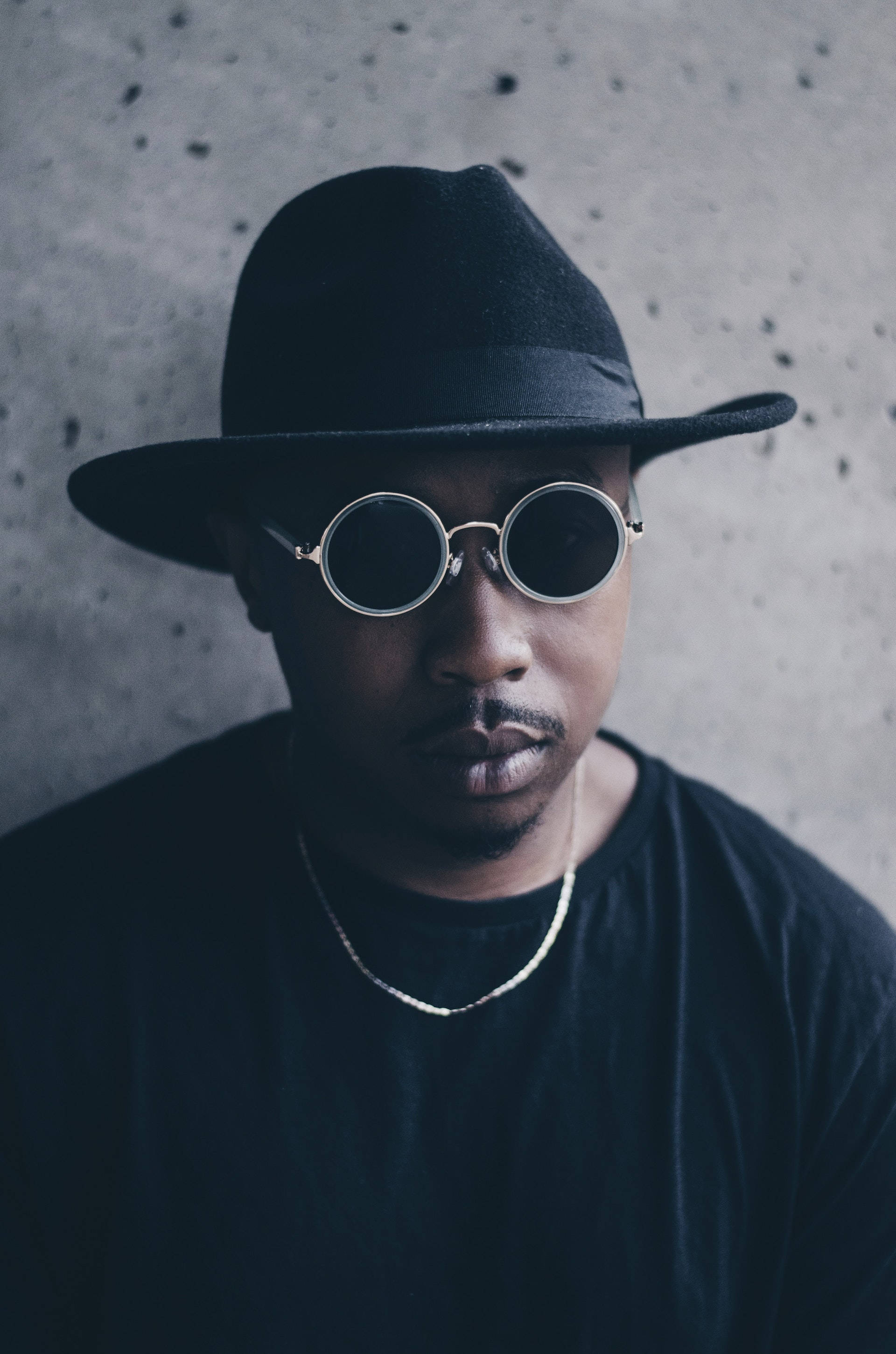 Sunglasses 桌布 壁紙 — 一個戴黑色軟呢帽和圓形太陽眼鏡的男人，對著有紋理的混凝土牆凝視。