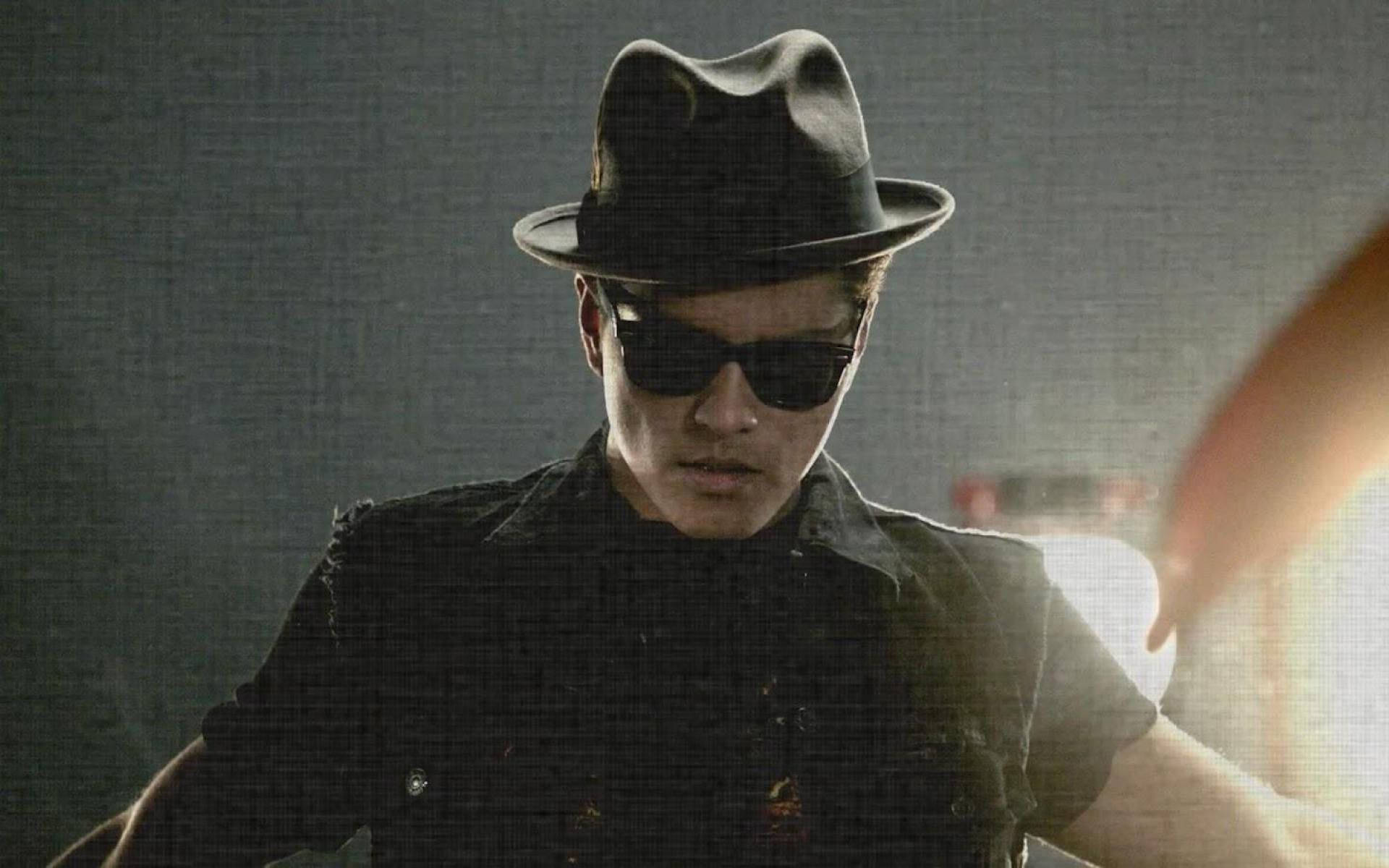 Bruno Mars 桌布 壁紙 — 一個戴黑色軟呢帽和太陽眼鏡的男人，在粗糙紋理背景和戲劇性背光下凝視著。