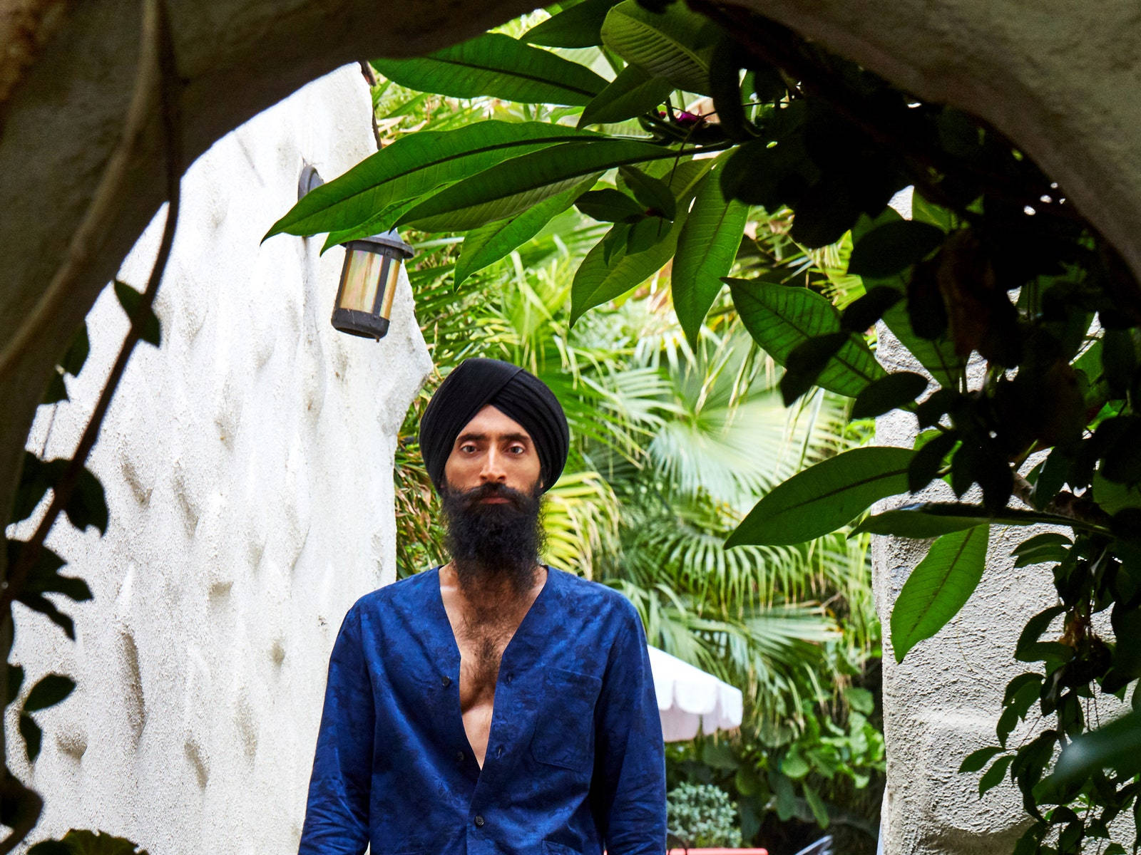 Garden 桌布 壁紙 — 一個戴黑色頭巾、留鬍子的男人，站在茂密的綠葉和紋理白色拱門之間。