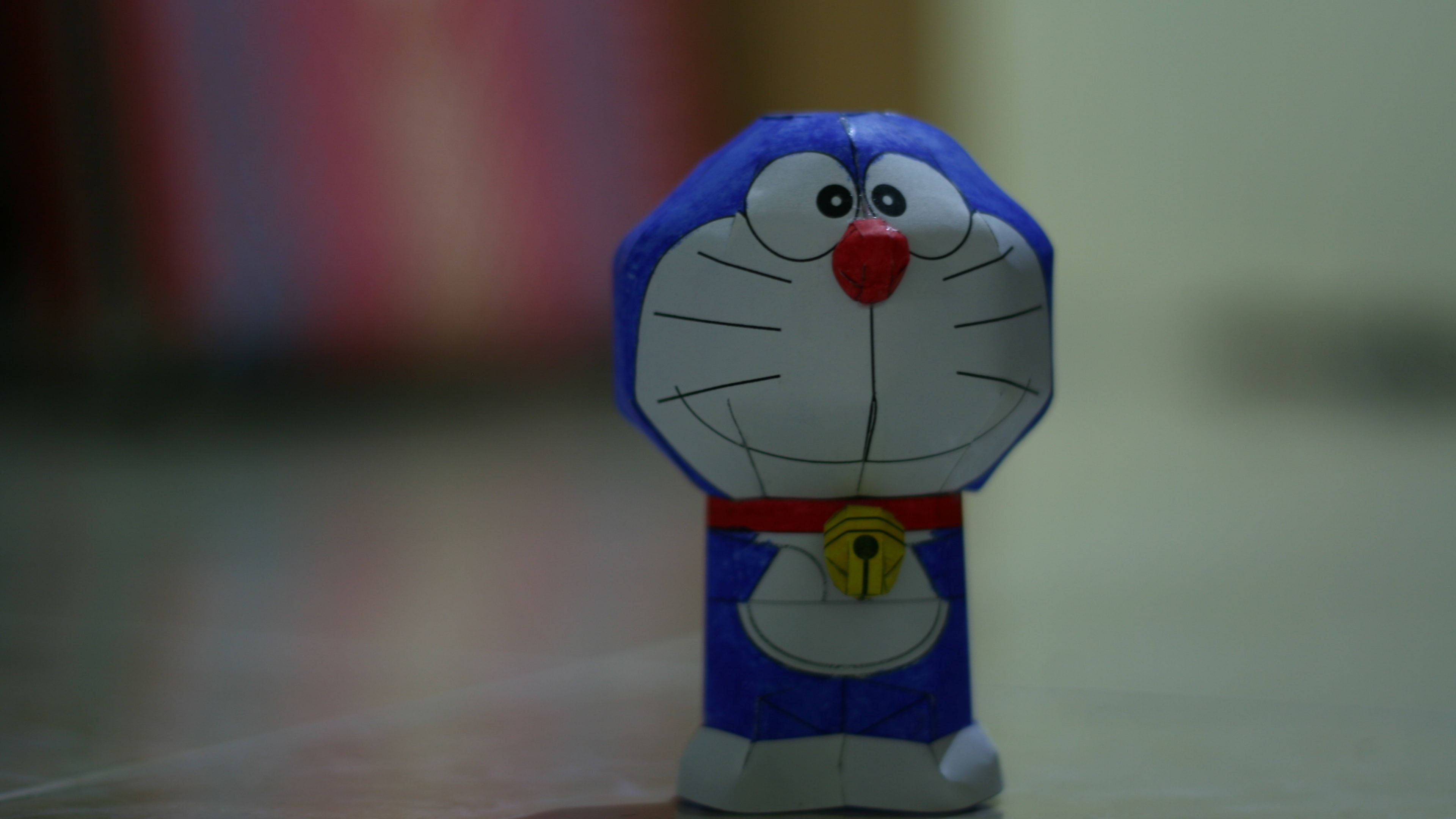 Doraemon 桌布 壁紙 — 一個手工紙製的哆啦A夢公仔，以柔和焦點站立，背景模糊色彩繽紛。