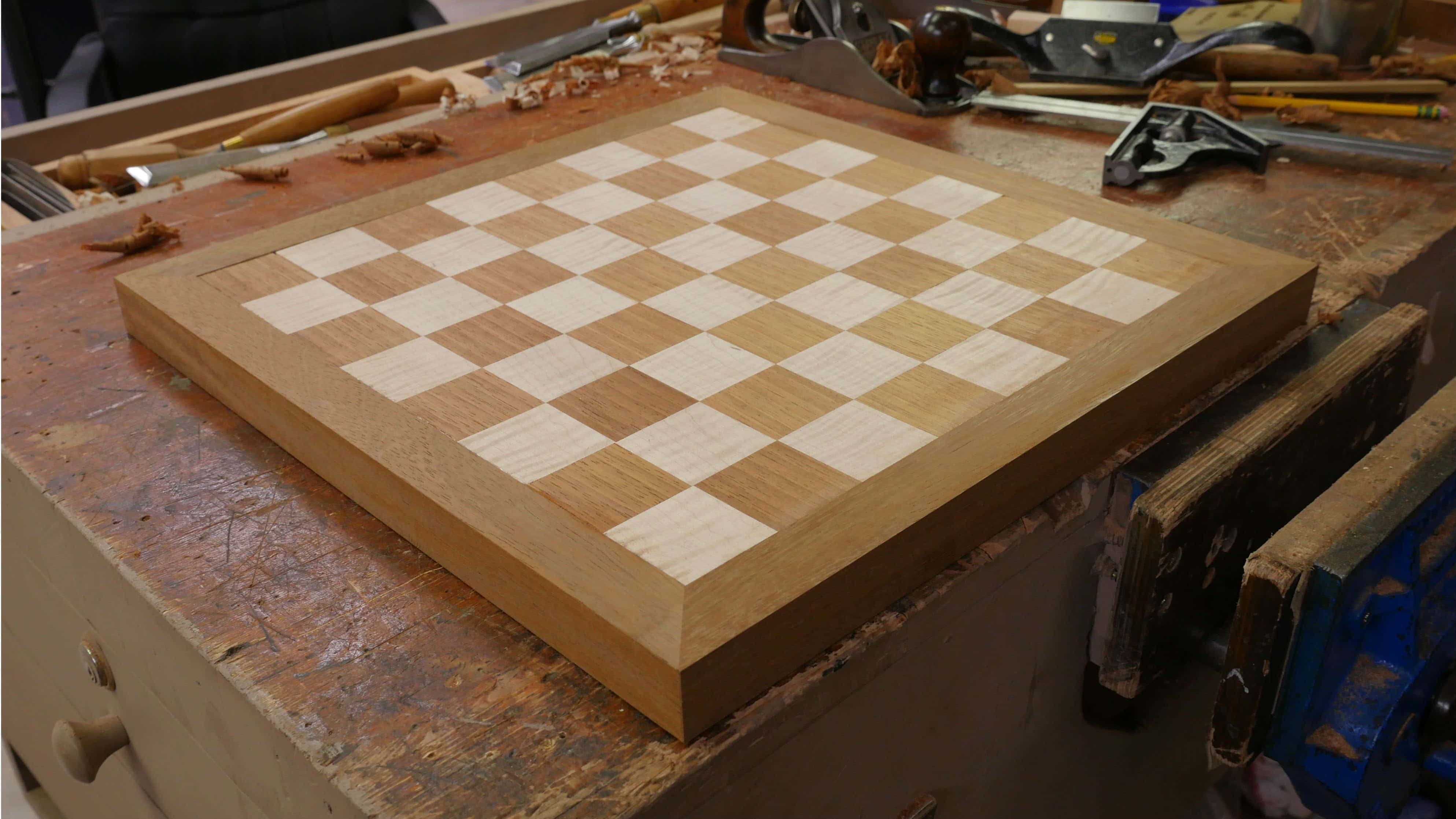 Chessboard 桌布 壁紙 — 一個手工製作的木製西洋棋棋盤，帶有淺色和深色橡木方格，放置在雜亂的木匠工作台上，周圍環繞著工具。