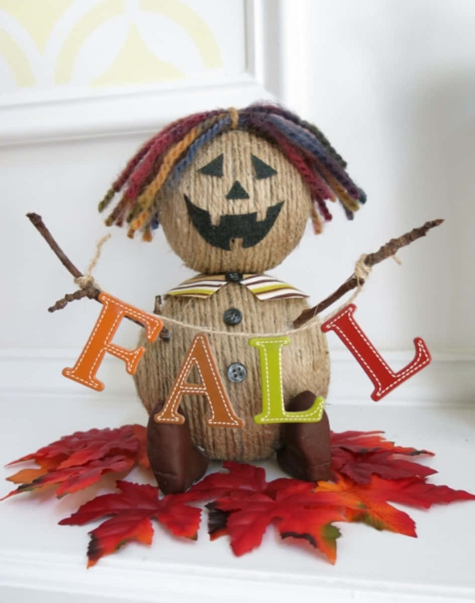Fall Scarecrow 桌布 壁紙 — 一個手工製作的毛線稻草人，帶有彩色辮子和南瓜燈臉，手持被紅楓葉包圍的「FALL」橫幅。