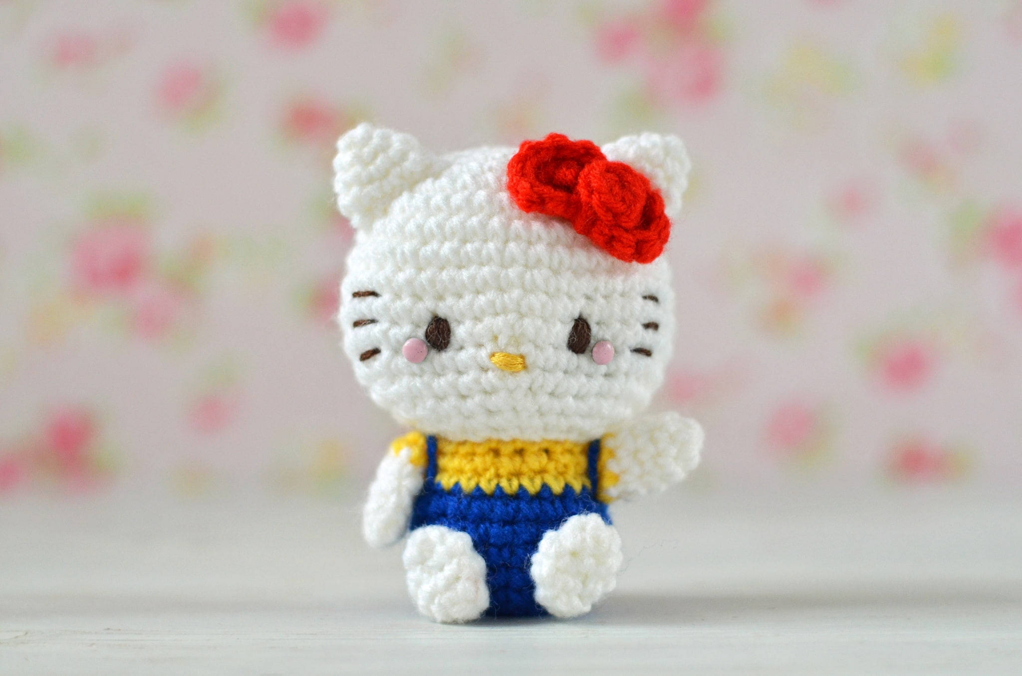 Hello 桌布 壁紙 — 一個手工製作的白色鉤針編織Hello Kitty公仔，戴著紅色蝴蝶結和藍色吊帶褲，坐在柔和的花卉背景前。