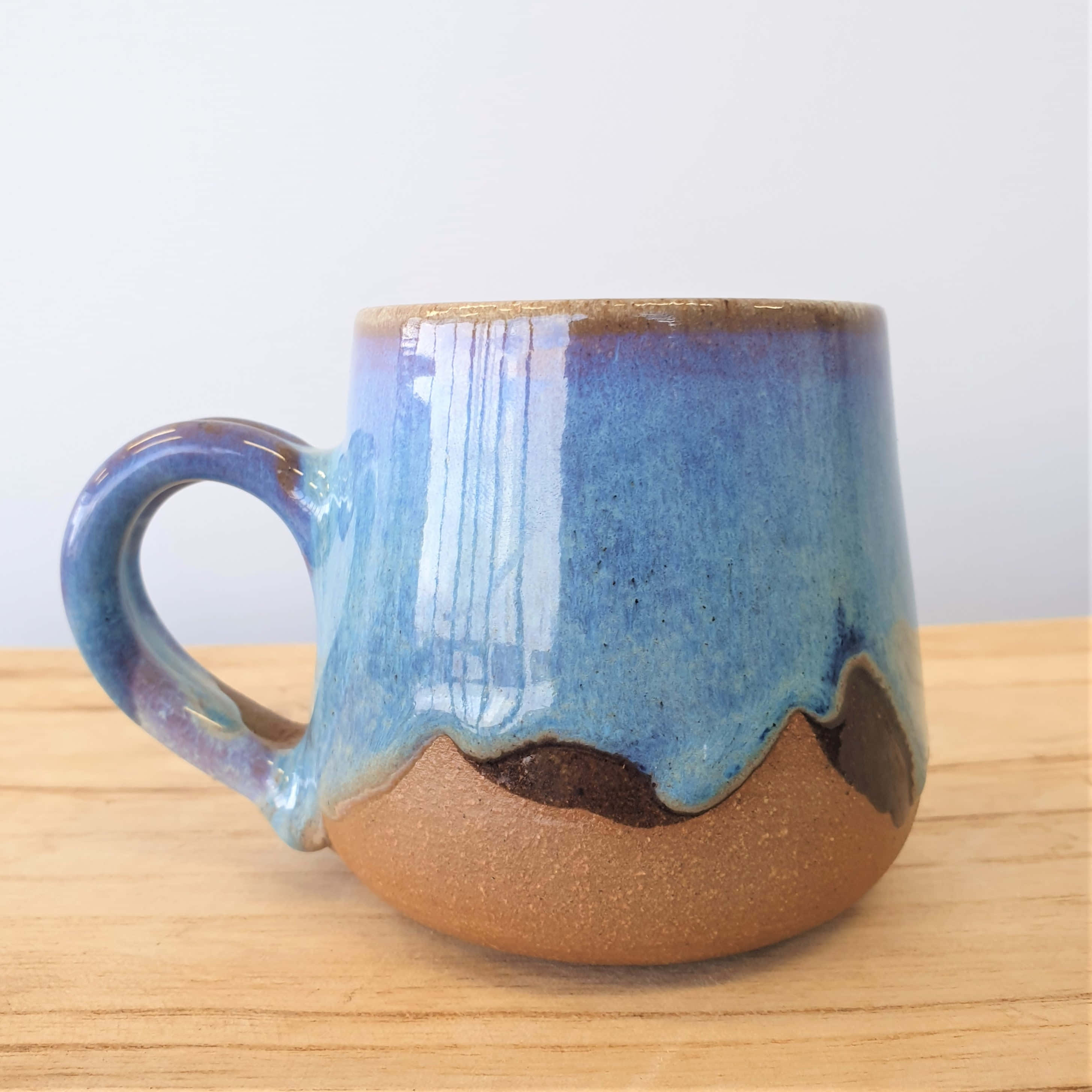 Ceramic 桌布 壁紙 — 一個手工製作的陶瓷杯，藍色釉料滴落在沙棕色底座上，放置在淺色木質表面上。