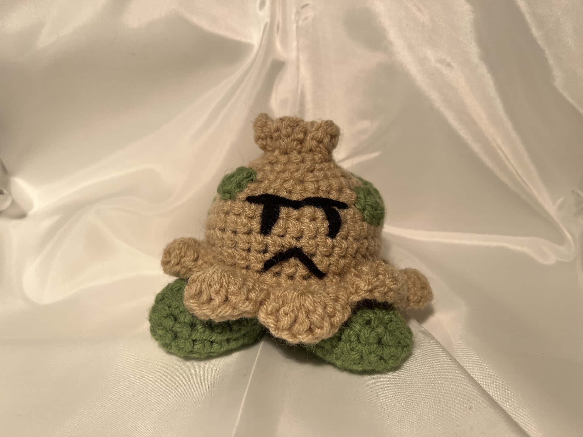 可愛 桌布 壁紙 — 一個手工鉤織的 Bulbasaur 毛絨玩具，擁有棕褐色身體和綠色葉子四肢，坐在白色緞面背景上。