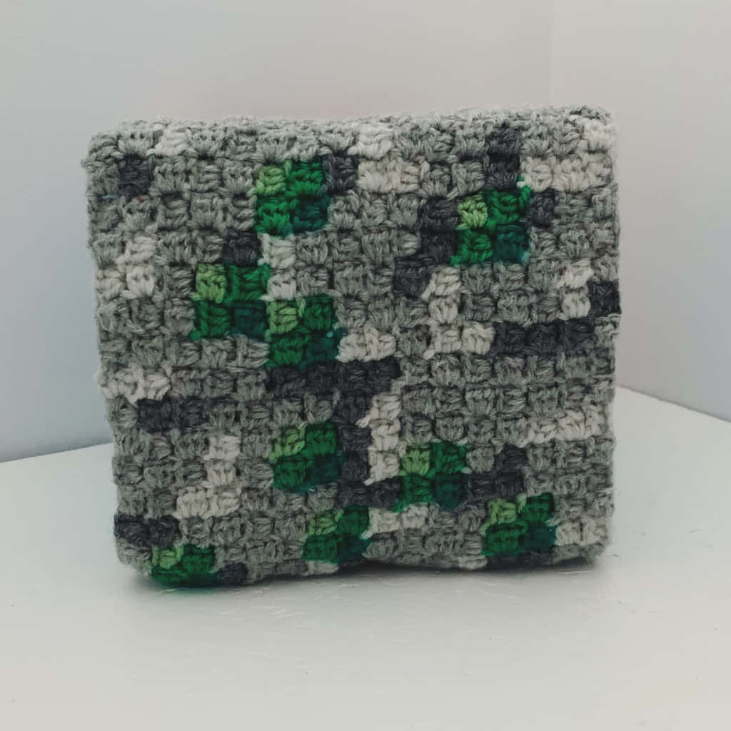 Minecraft Emeralds 桌布 壁紙 — 一個手工鉤織的正方形，帶有像素化的綠色和灰色迷彩圖案，放在白色表面上。