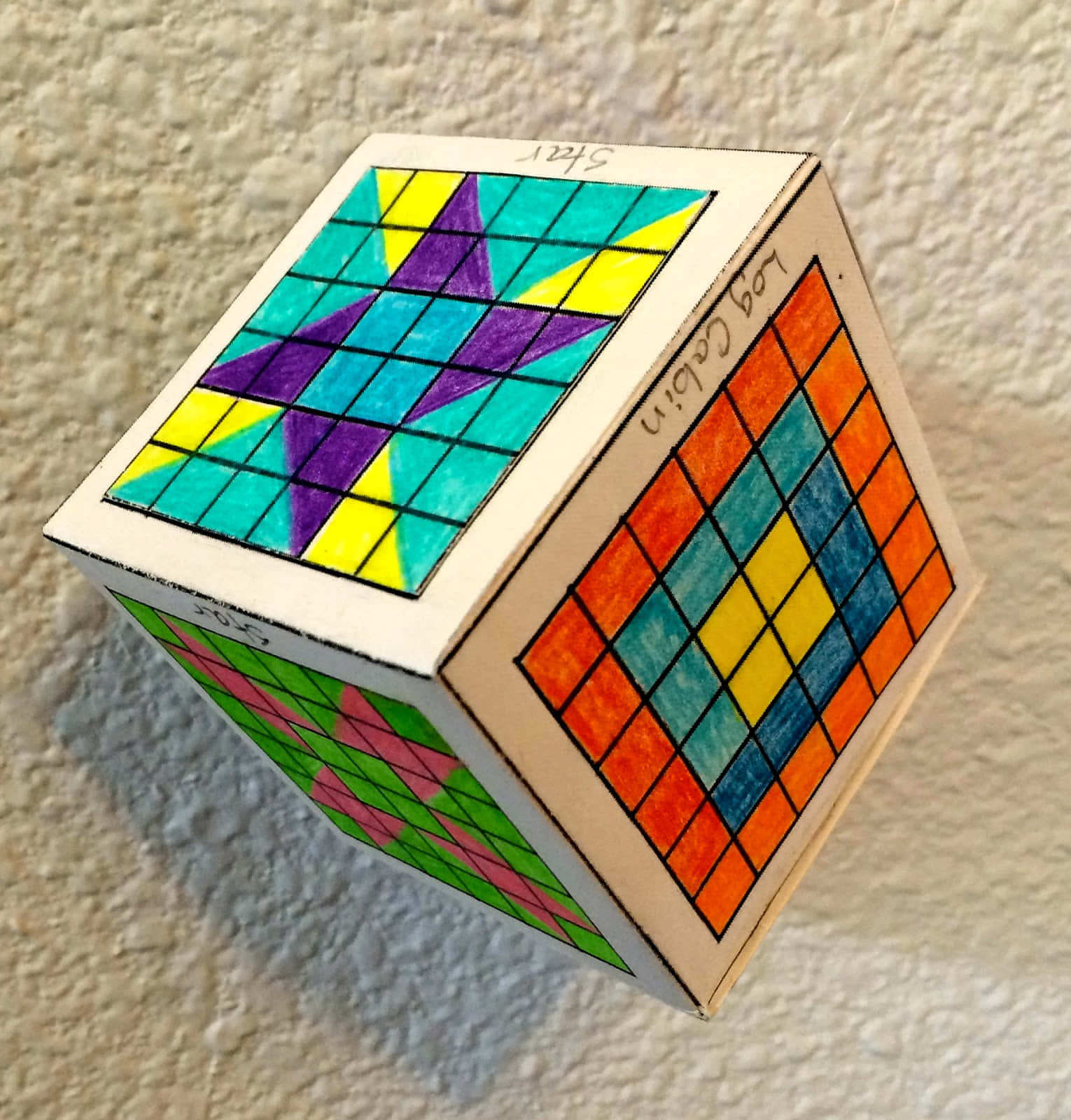 Block 桌布 壁紙 — 一個手繪彩色木立方體，帶有幾何圖案和手寫標籤，放置在紋理牆壁上。