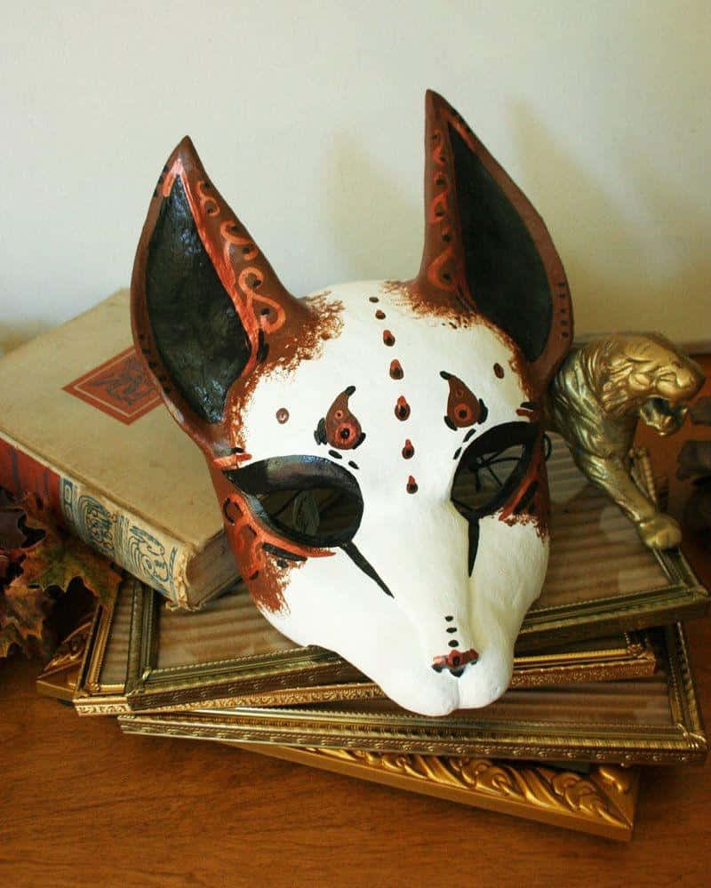 Yokai 桌布 壁紙 — 一個手繪狐狸面具，白色底色搭配橙色黑色部落圖案，放置在華麗的金色畫框和一本舊書上。