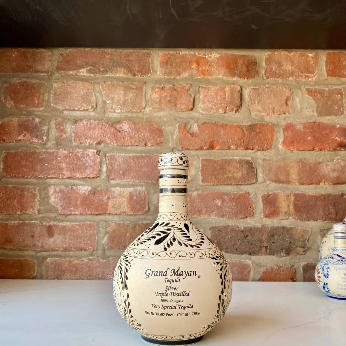 alcohol 桌布 壁紙 — 一個手繪的Grand Mayan龍舌蘭酒瓶，帶有黑色花卉圖案，放在白色檯面上，背景是質樸的紅磚牆。