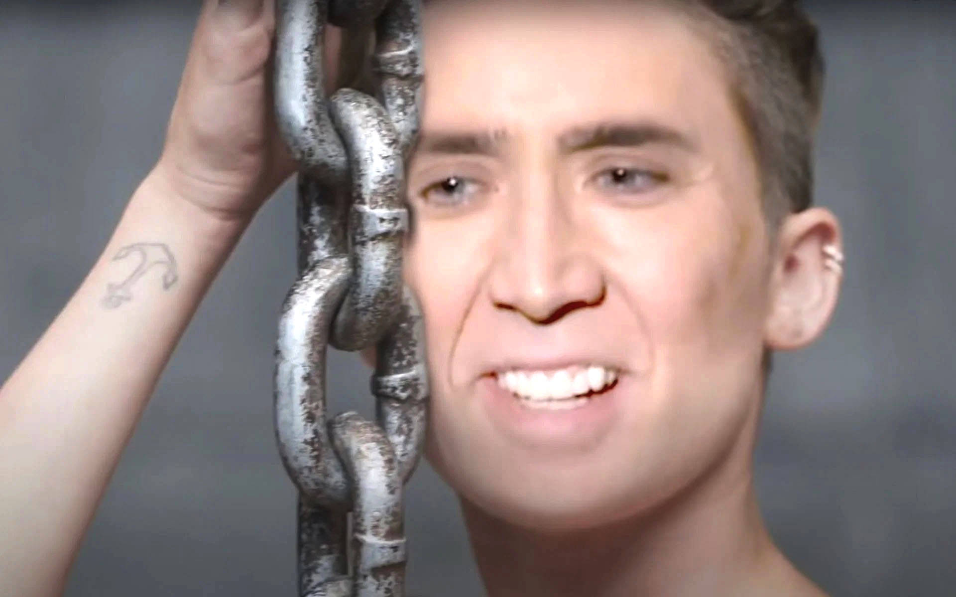 nicolas cage meme 桌布 壁紙 — 一個手腕上有錨紋身的男人，在灰色背景前抓著一條沉重的生鏽鐵鏈。