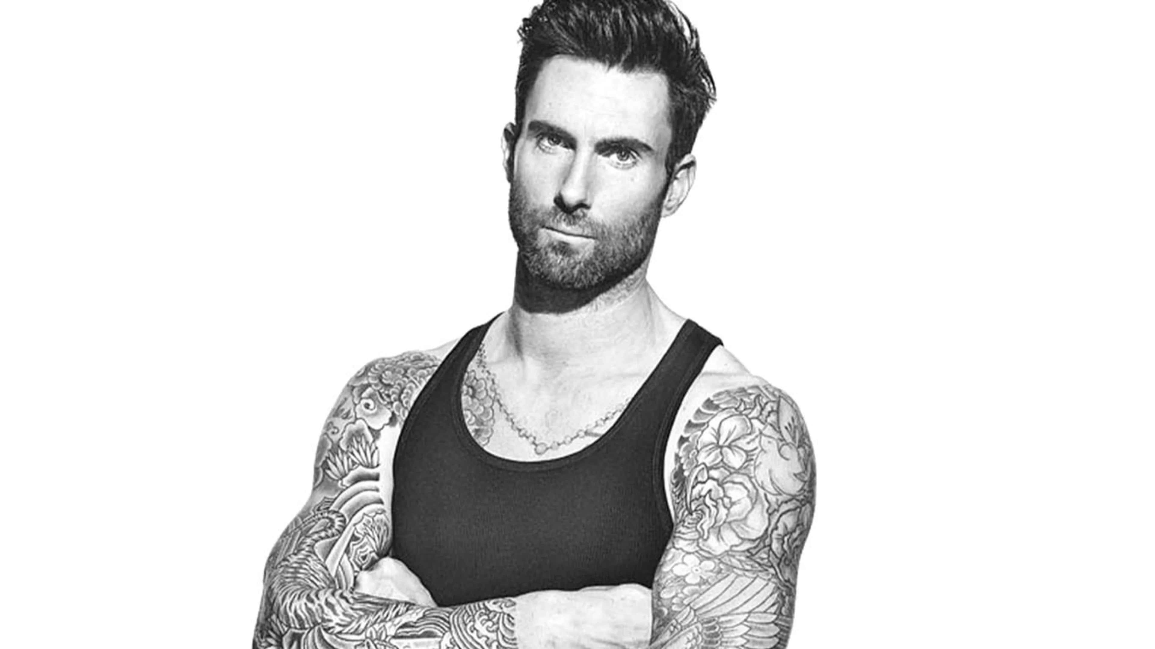 Adam Levine 桌布 壁紙 — 一個手臂和胸口有刺青的男人，穿著黑色背心，在白色背景前自信地凝視鏡頭。