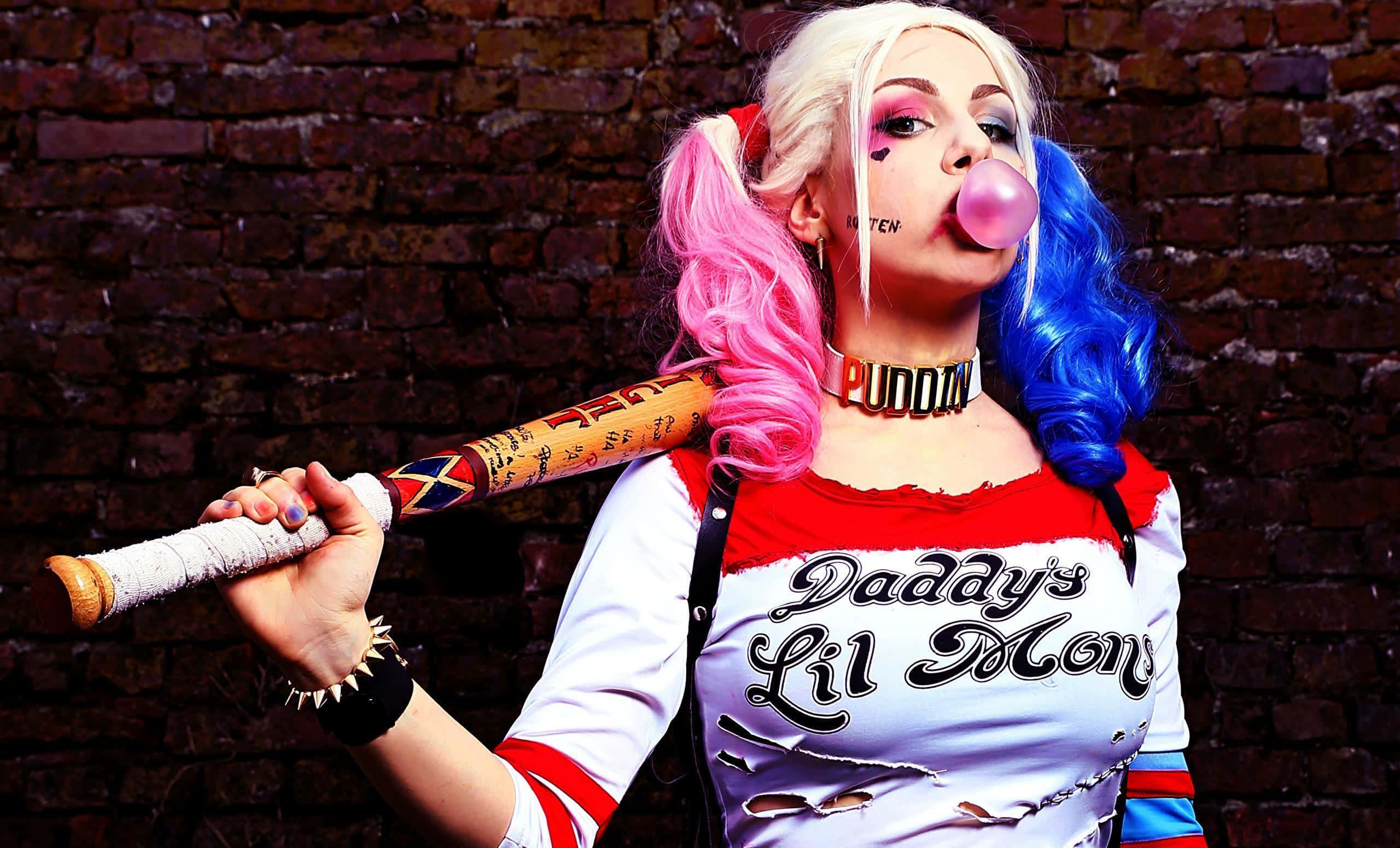 Harley Quinn Baseball Bat 桌布 壁紙 — 一個扮演哈莉·奎茵的人，一邊對著磚牆拿著棒球棍，一邊吹著口香糖泡泡。