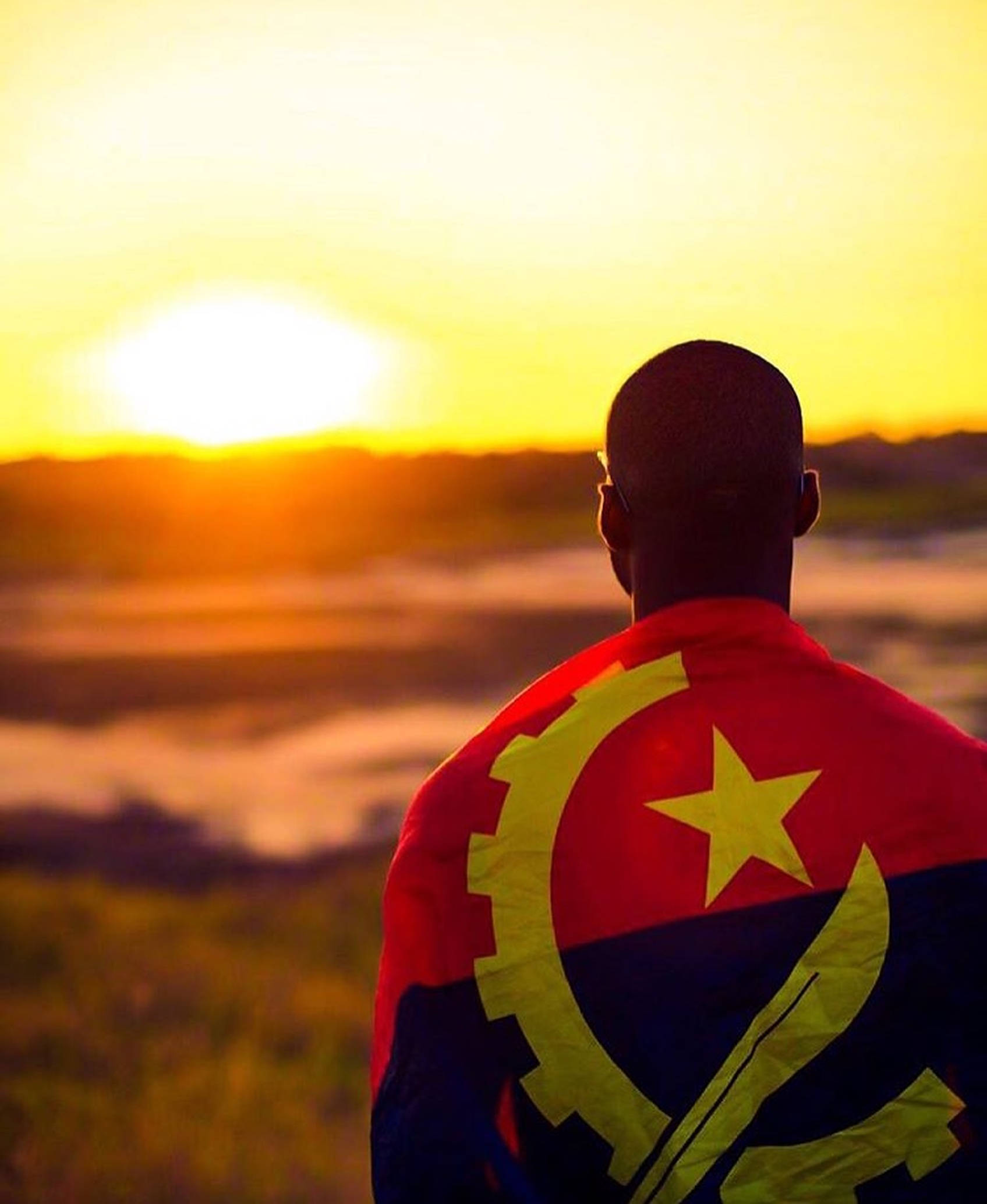 Angola 桌布 壁紙 — 一個披著安哥拉國旗的人，在霧氣瀰漫的田野上，以金色日出為背景形成剪影。