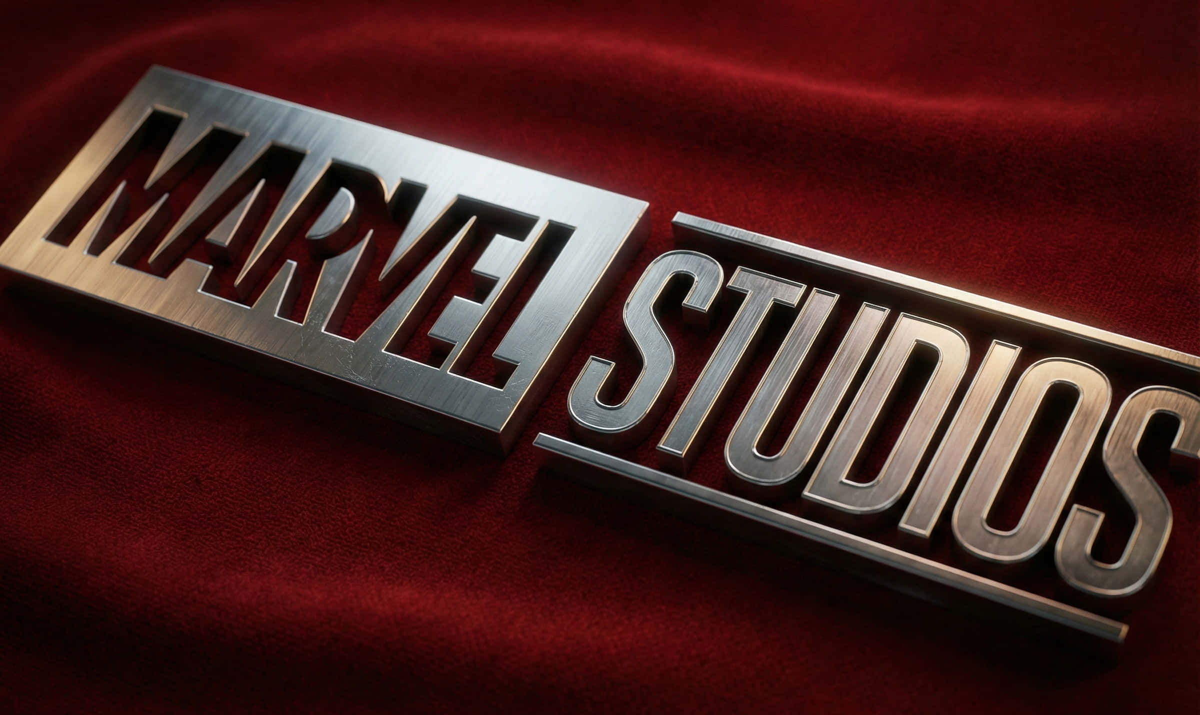 Mcu 桌布 壁紙 — 一個拉絲金屬Marvel Studios標誌對角放置在豐富的紅色天鵝絨表面上，帶有戲劇性的燈光。