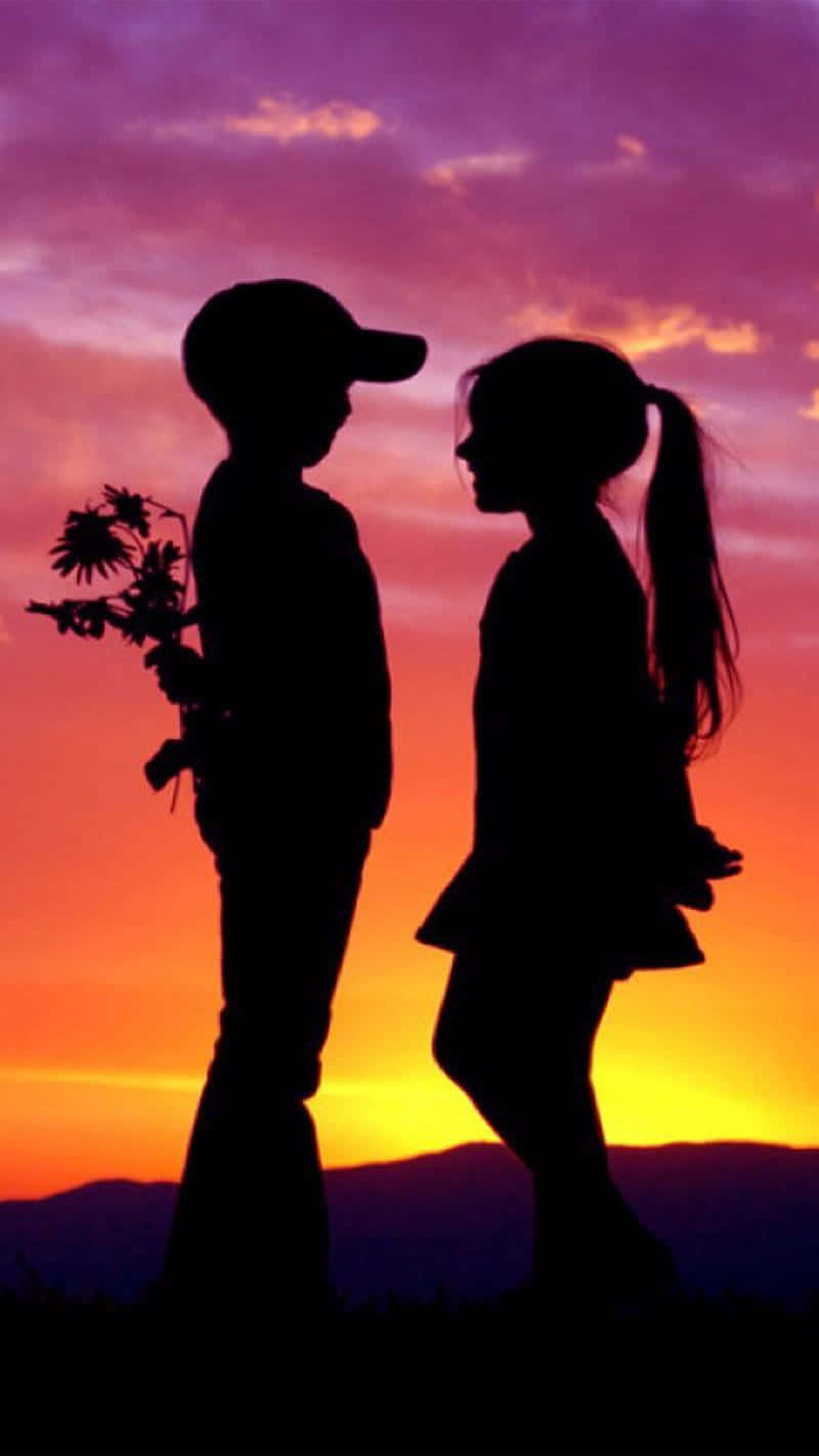 Silhouette 桌布 壁紙 — 一個拿著向日葵的男孩和一個紮馬尾的女孩的剪影，在充滿活力的日落天空下相對而立。