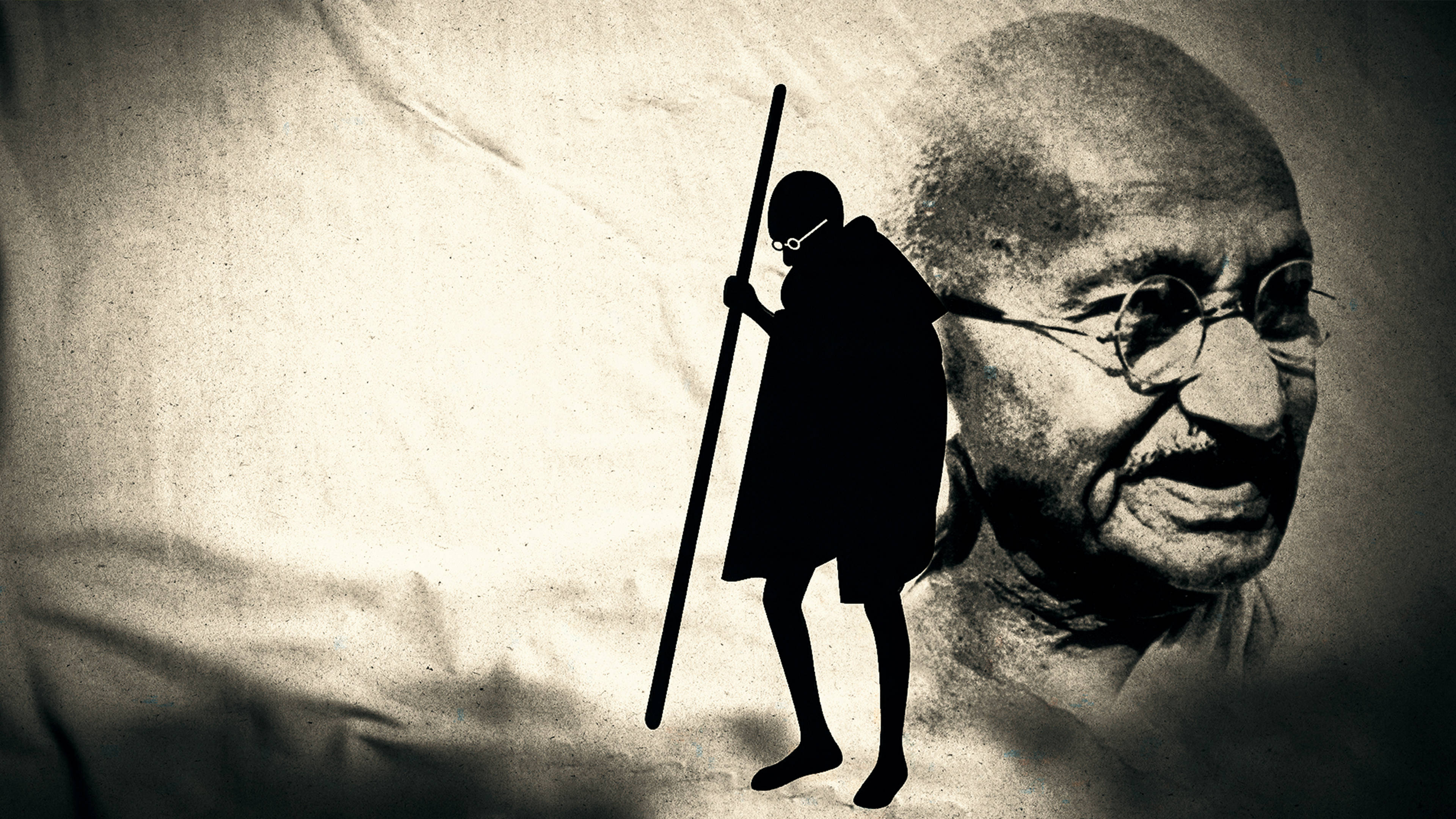 Mahatma Gandhi 桌布 壁紙 — 一個拿著手杖行走的人物的鮮明黑色剪影，站在一幅大型的深褐色調的聖雄甘地肖像旁邊。