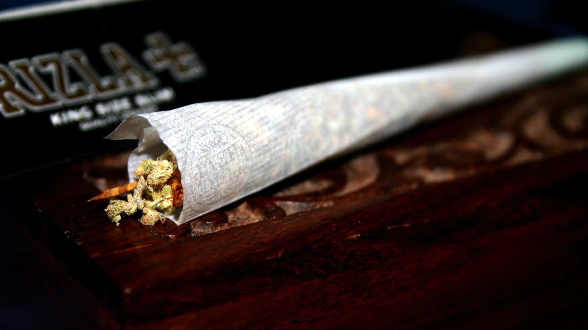 大麻 桌布 壁紙 — 一個捲好的大麻煙捲特寫，放在深色木質表面上，背景有一個品牌盒子。