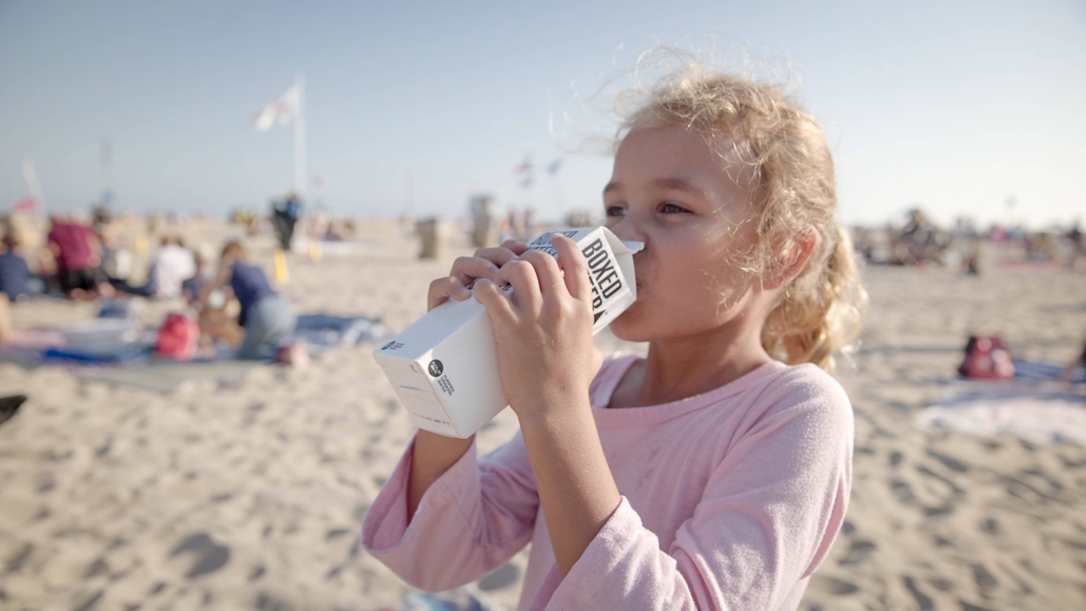 Drink Water 桌布 壁紙 — 一個捲曲金髮的小女孩在陽光明媚的海灘上喝著白色盒裝飲料，背景中人們在放鬆。