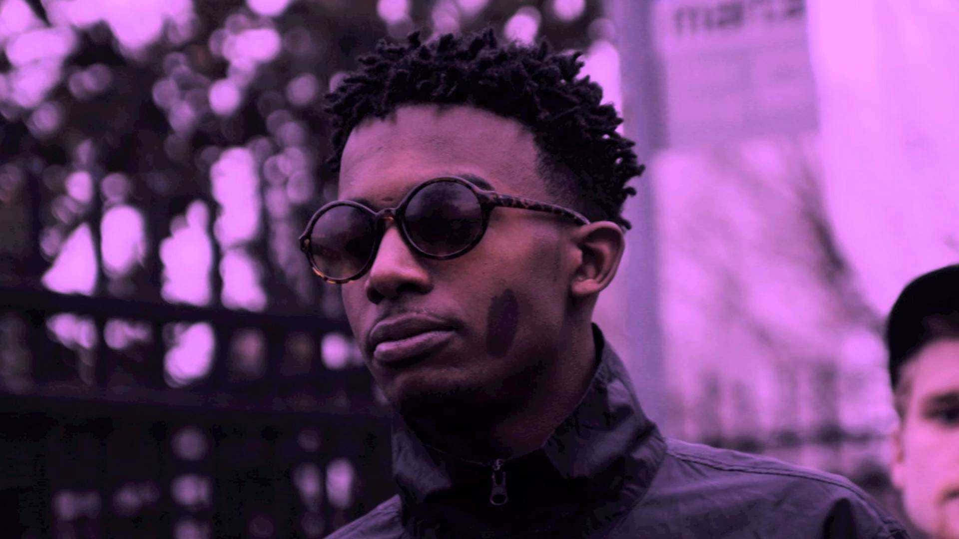 Playboi Carti 桌布 壁紙 — 一個捲髮、戴圓形太陽眼鏡的男人，在紫色濾鏡和模糊的城市背景下，視線離開鏡頭。