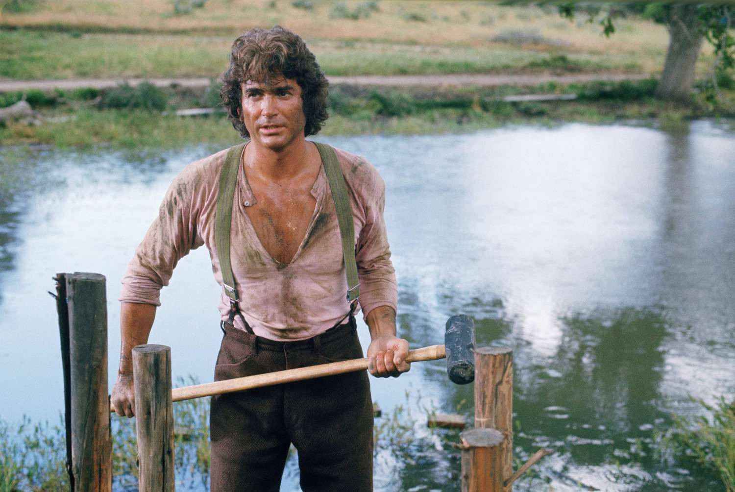 Michael Landon 桌布 壁紙 — 一個捲髮、衣服沾滿泥濘的男人站在河邊，在木樁之間拿著大錘。