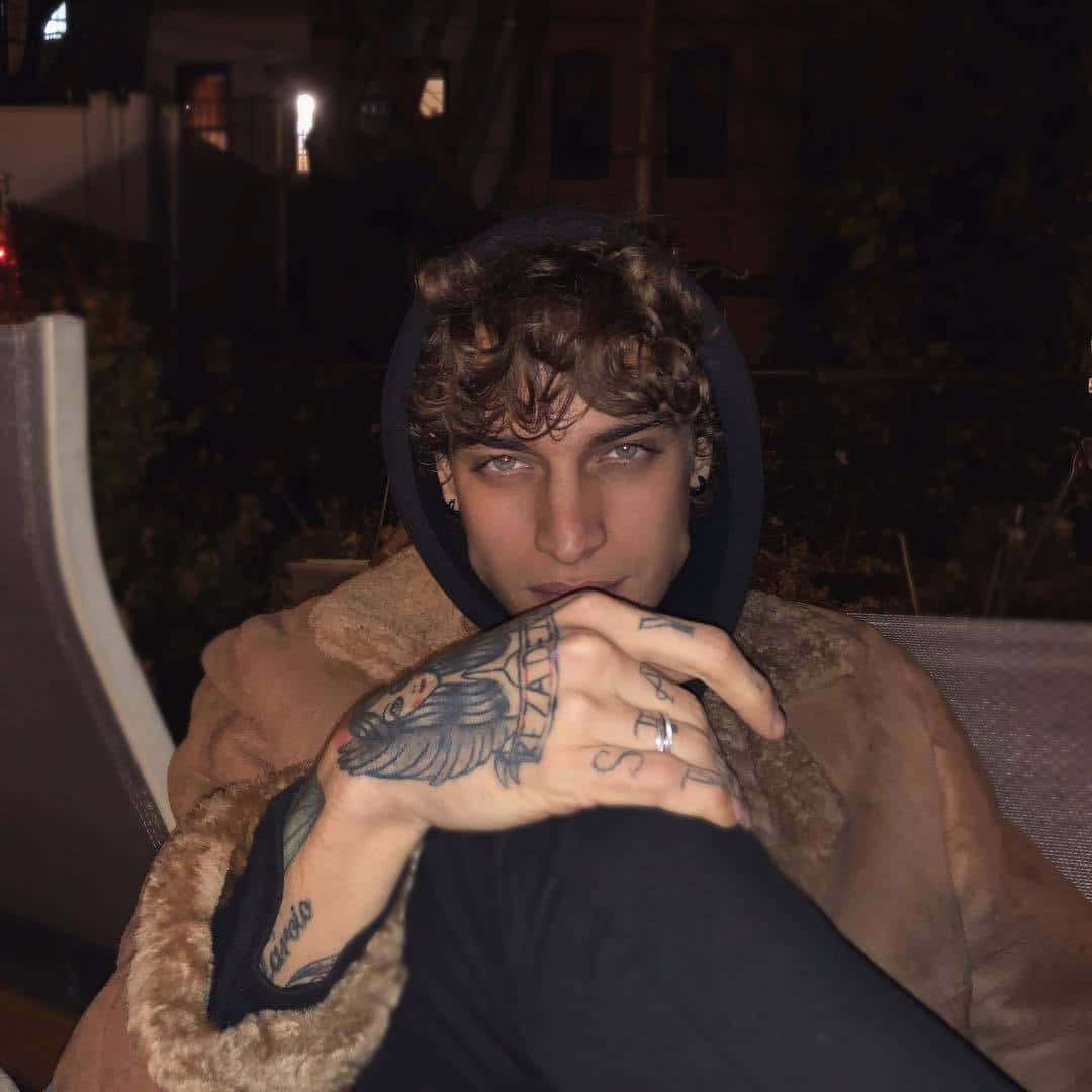 Tattoo Boy 桌布 壁紙 — 一個捲髮且有刺青的年輕男子，在夜晚將有刺青的手放在連帽衫下的臉旁休息。