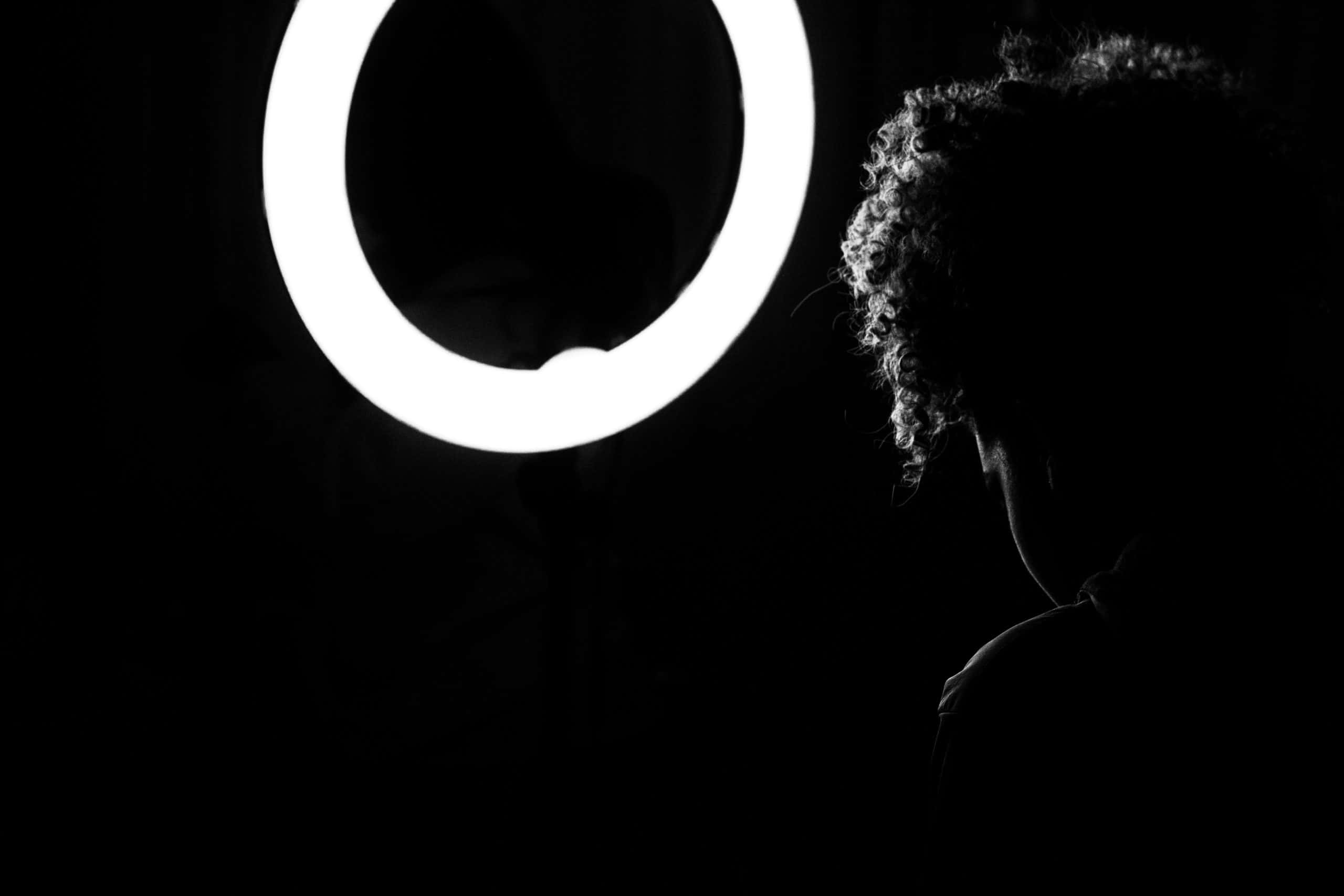 Ring Light 桌布 壁紙 — 一個捲髮人物的剪影，在漆黑背景中凝視著發光的環形燈。