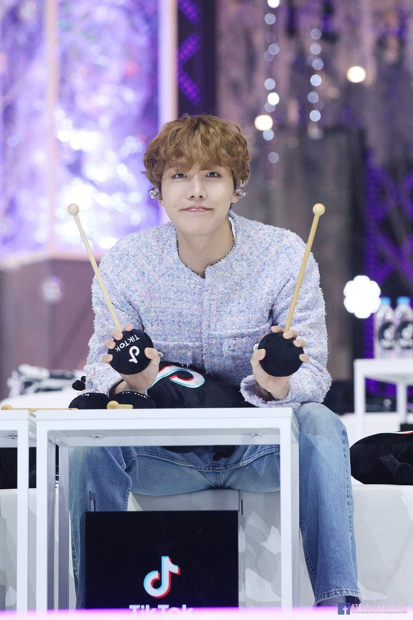 jhope 桌布 壁紙 — 一個捲髮年輕男子坐著，手持鼓棒和TikTok品牌的鼓面，背景是充滿活力的紫色散景。