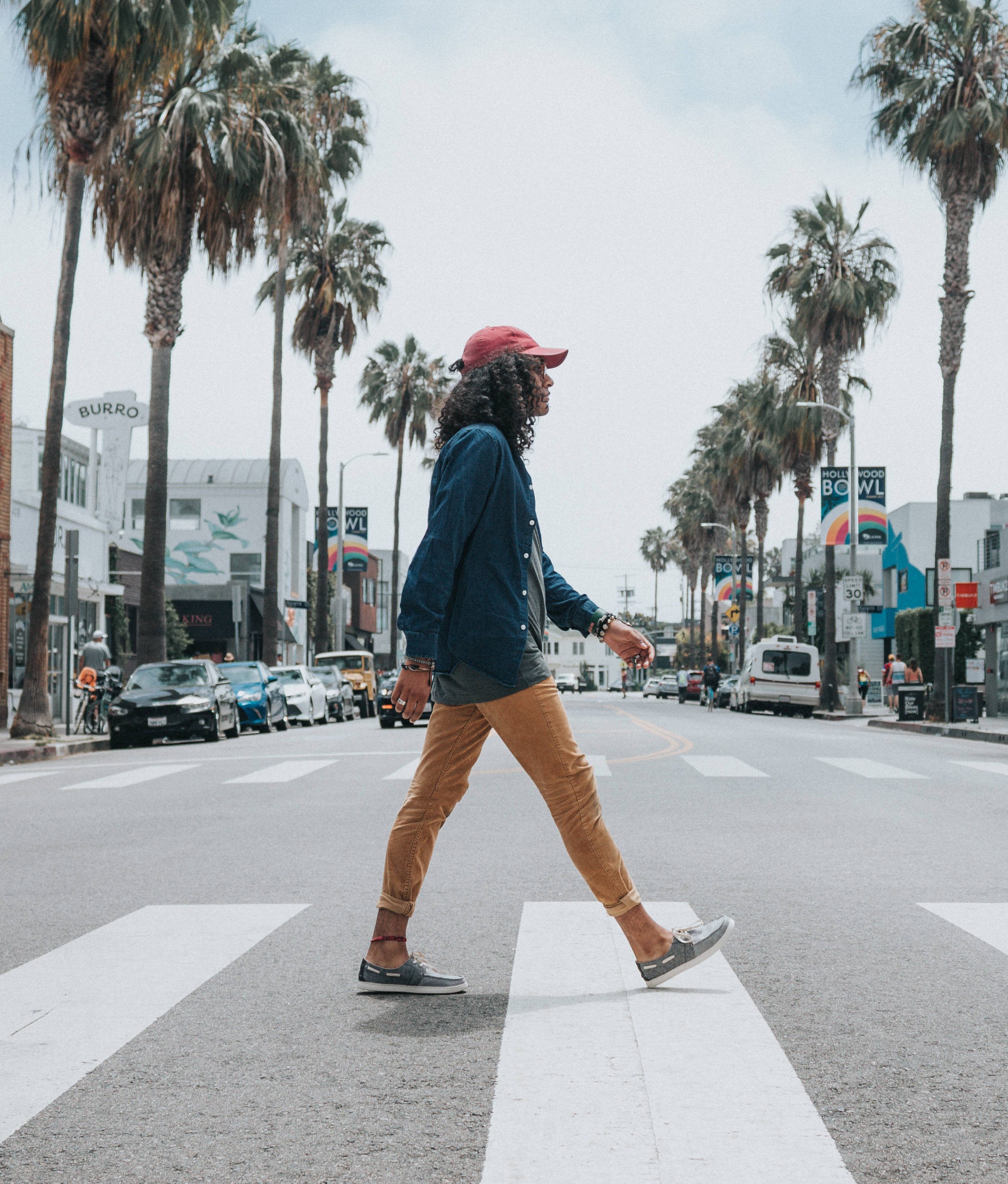 Trendy 桌布 壁紙 — 一個捲髮戴紅帽的人正在穿過一條兩旁種有高大棕櫚樹的街道，天空多雲。