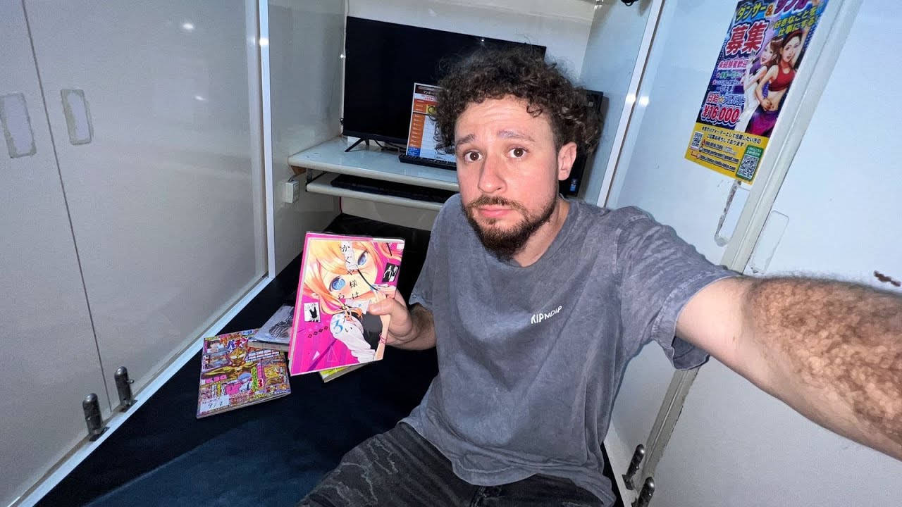 日本 桌布 壁紙 — 一個捲髮男人坐在一個狹小房間裡，拿著一本色彩繽紛的漫畫書自拍。