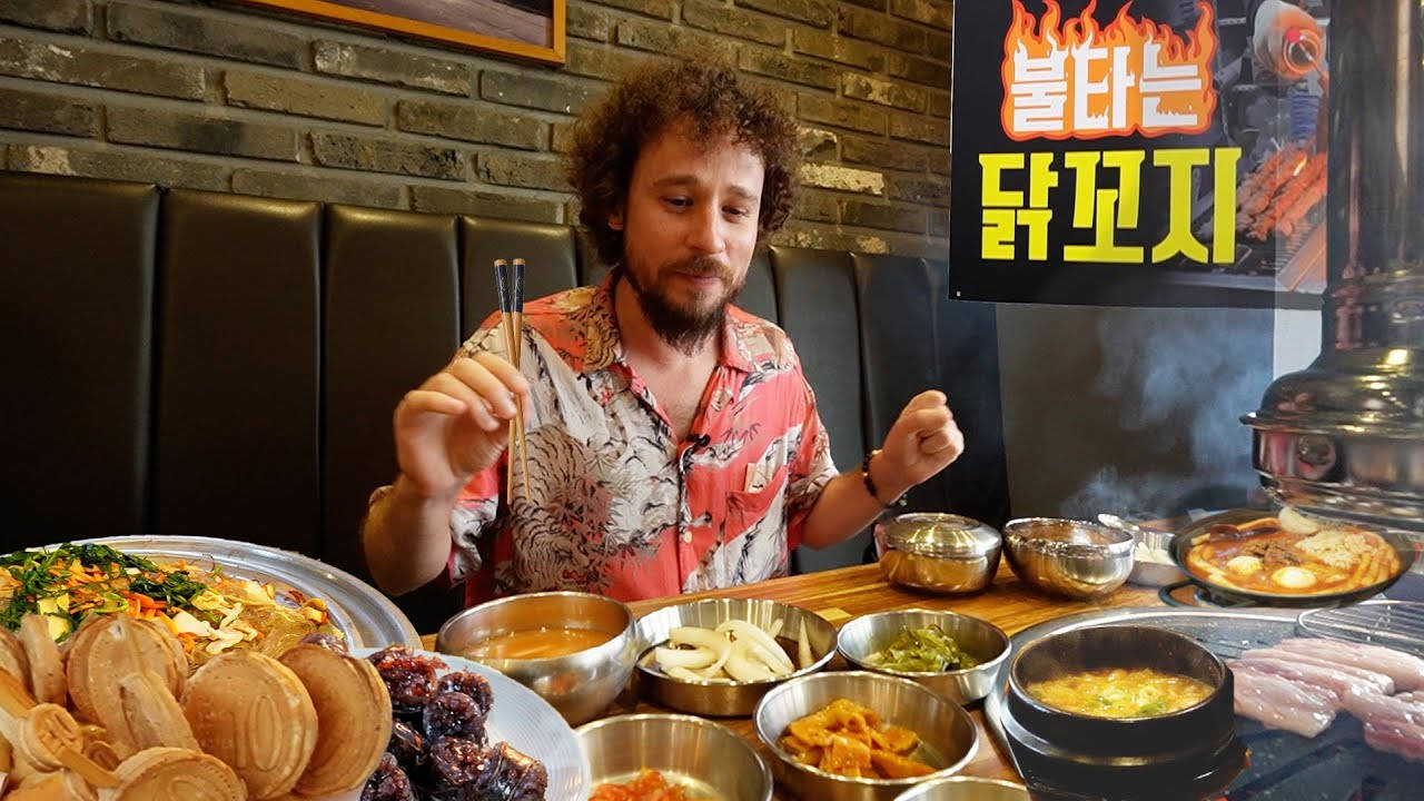 Eating 桌布 壁紙 — 一個捲髮男人坐在一張擺滿韓國燒烤和配菜的桌子旁，餐廳有磚牆。