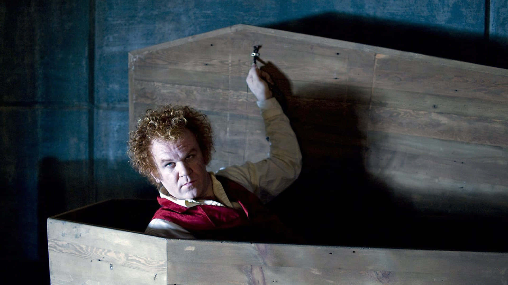 John C. Reilly 桌布 壁紙 — 一個捲髮男人坐在木棺內，手持一個小物件，專注地凝視著相機。