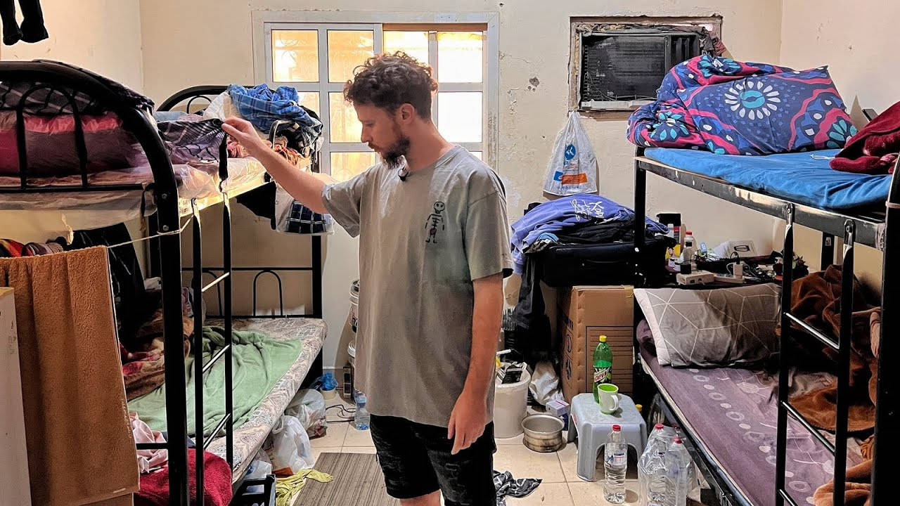 Bedroom 桌布 壁紙 — 一個捲髮男子站在雜亂的宿舍房間裡，房間裡有雙層床和散落的個人物品，在溫暖的室內燈光下。
