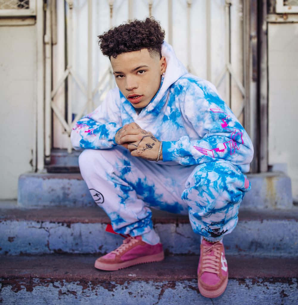 Lil Mosey 桌布 壁紙 — 一個捲髮的年輕人蹲在風化的階梯上，穿著藍色紮染連帽衫和粉紅色運動鞋。