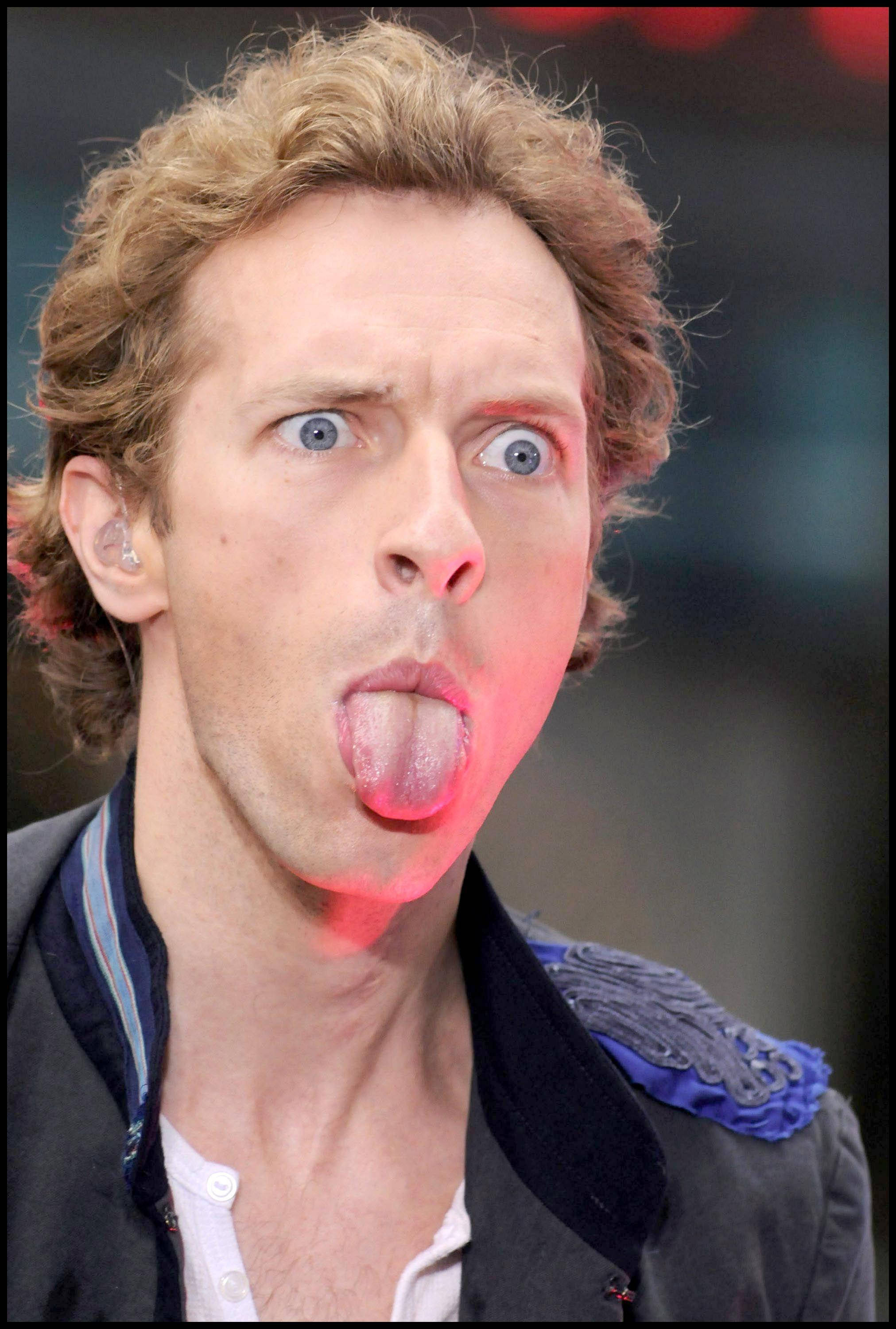 Chris Martin 桌布 壁紙 — 一個捲髮藍眼的男人在舞台燈光下做出頑皮的表情伸出舌頭。