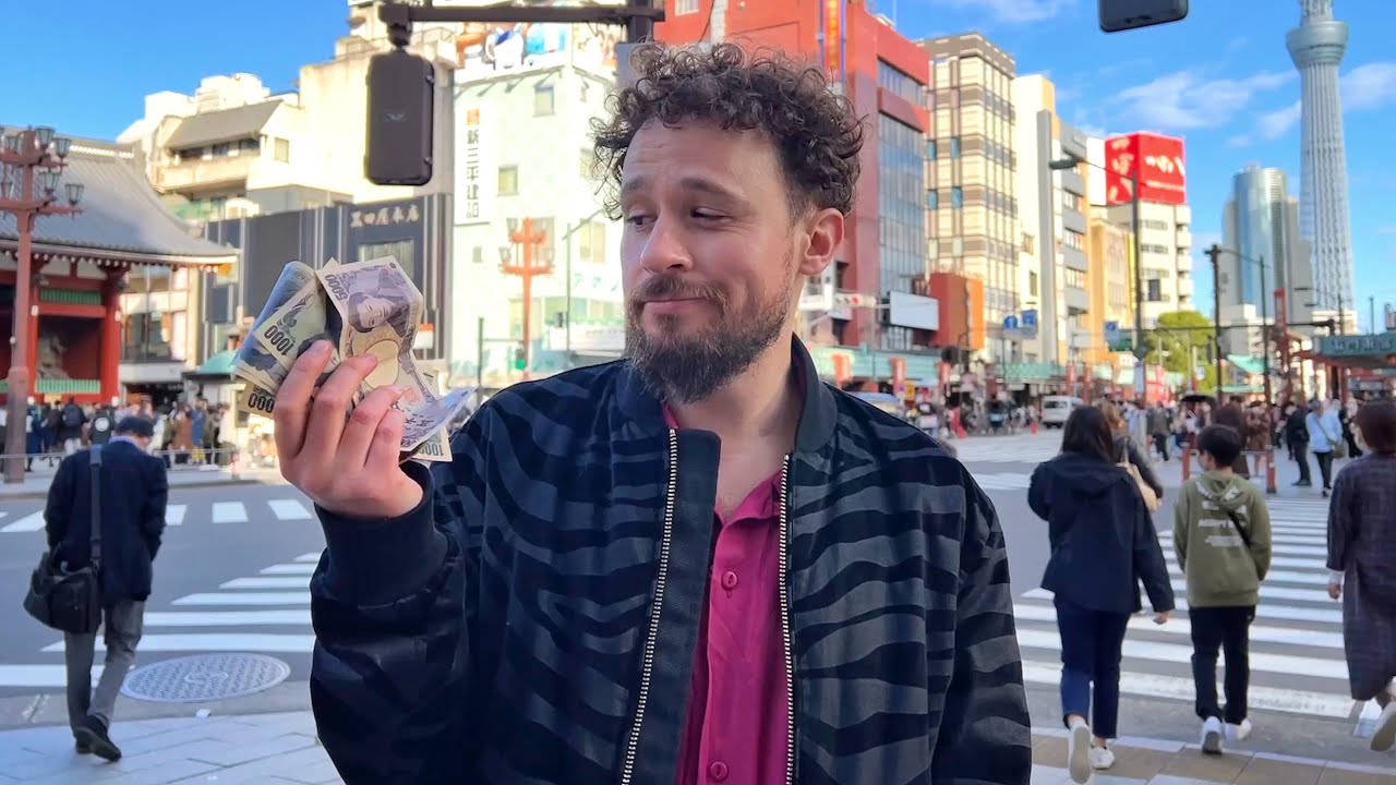 東京 桌布 壁紙 — 一個捲髮鬍鬚男子站在繁忙的東京街道上，背景可見東京晴空塔，手持日本日元紙幣。