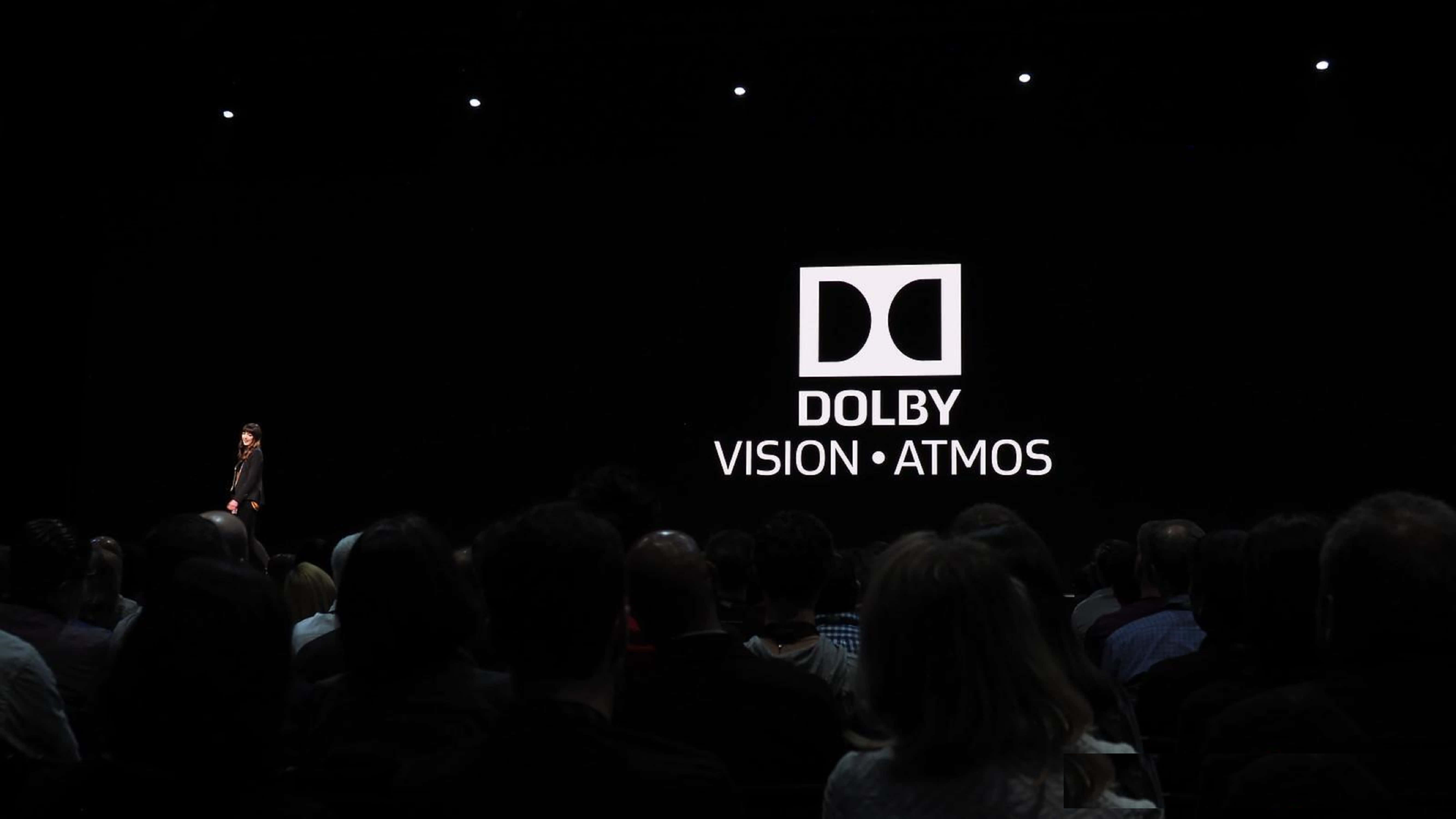 4K Dolby Vision 桌布 壁紙 — 一個揚聲器獨自站在黑暗的舞台上，面前是一個顯示Dolby Vision Atmos標誌的大螢幕，觀眾坐在昏暗的燈光中。