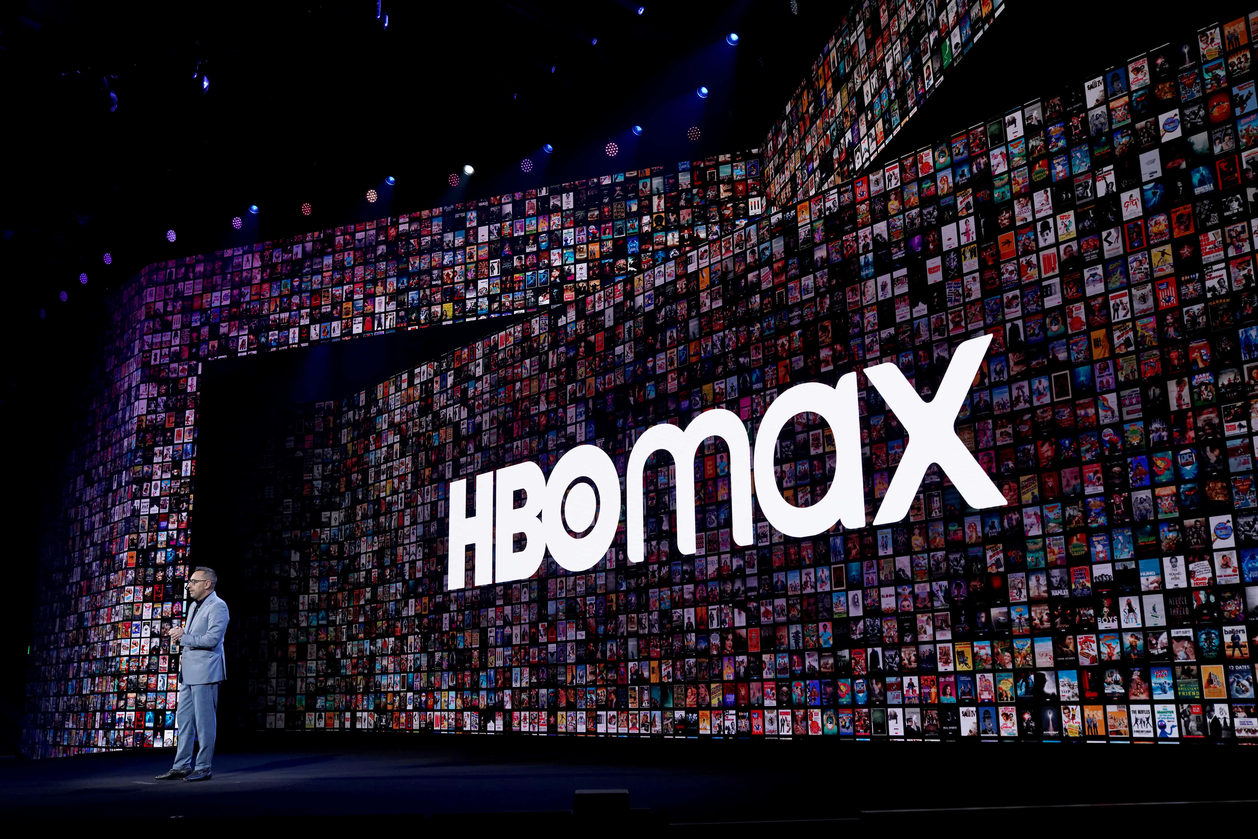 HBO 桌布 壁紙 — 一個揚聲器站在黑暗的舞台上，面前是一幅巨大的彩色電影海報牆，由聚光燈照亮，並帶有發光的HBO Max標誌。