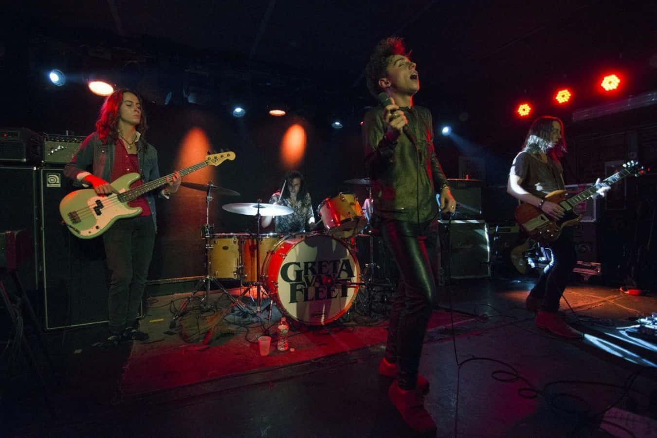 band 桌布 壁紙 — 一個搖滾樂團在昏暗的舞台上充滿活力地表演，舞台被紅色和橙色的聚光燈籠罩，鼓手在 'Greta Van Fleet' 鼓組後面。