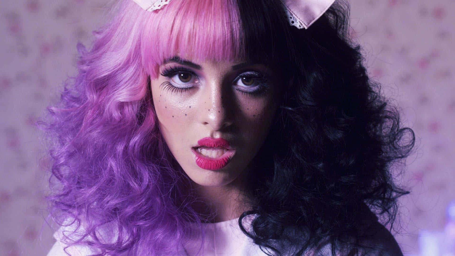Melanie Martinez Aesthetic 桌布 壁紙 — 一個擁有一半粉紅一半黑色捲髮、化著大膽妝容的人，在花卉壁紙背景下強烈地凝視著。