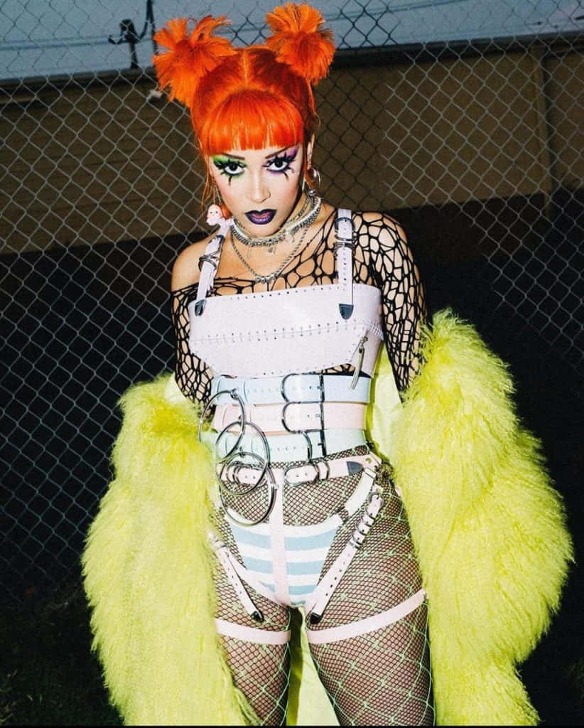 Doja Cat 桌布 壁紙 — 一個擁有亮橙色辮子和誇張妝容的人，穿著白色束帶服裝，搭配螢光黃色毛絨袖子，站在鐵絲網圍欄前。