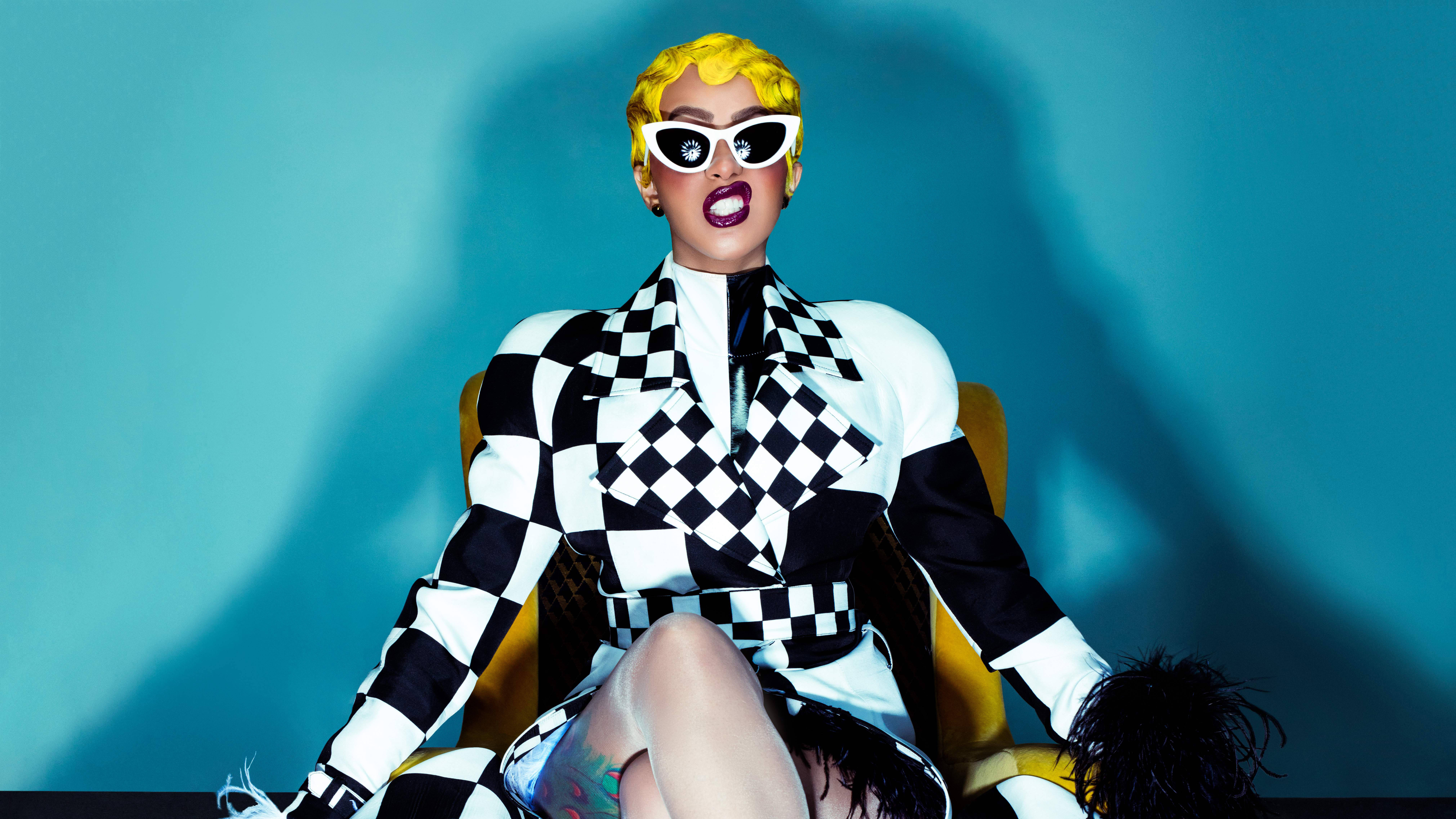 cardi 桌布 壁紙 — 一個擁有亮麗黃色頭髮和白色太陽眼鏡的人，自信地坐在黃色椅子上，穿著大膽的黑白格紋服裝，背景是藍綠色。