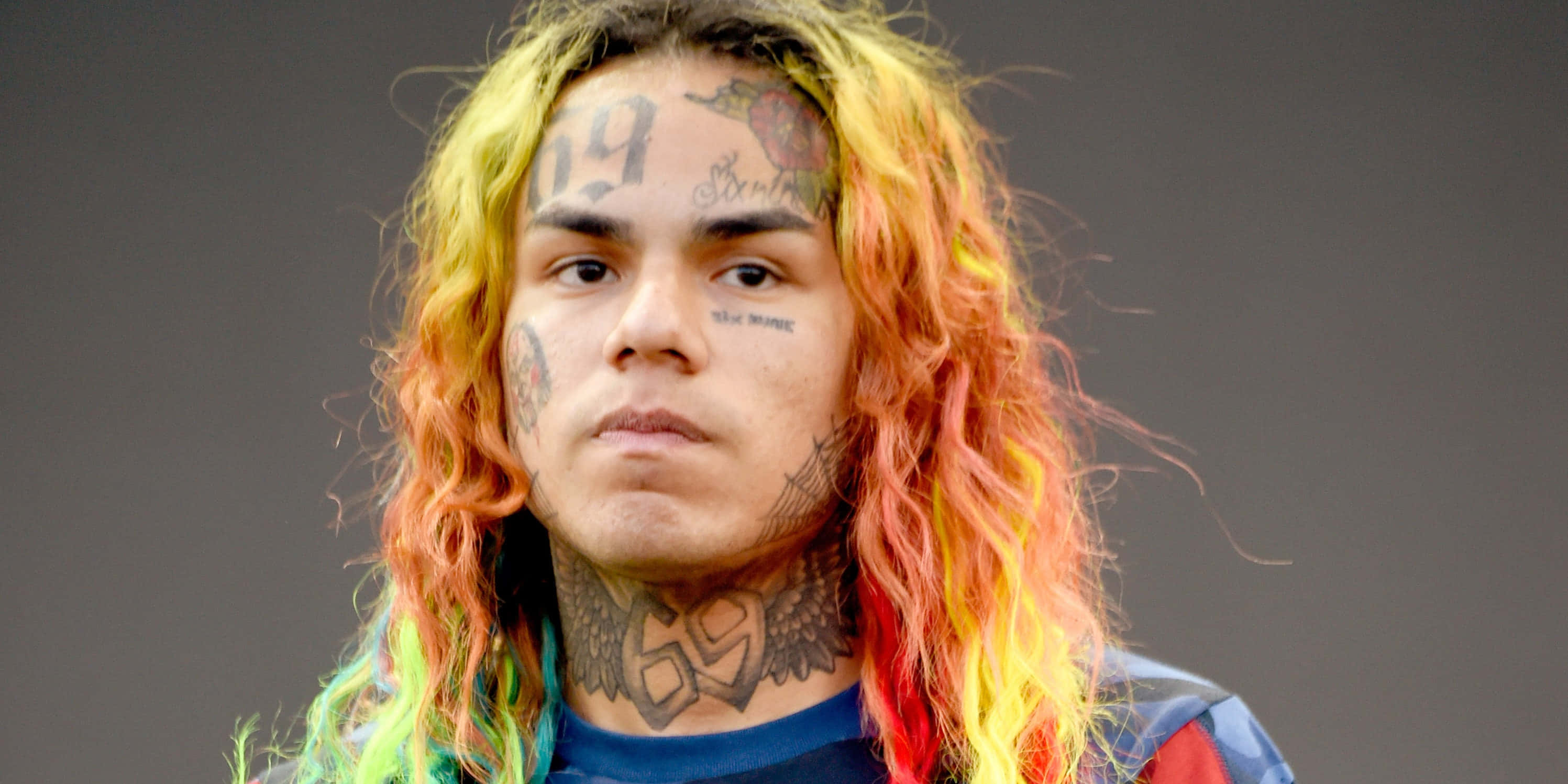 69 Rapper 桌布 壁紙 — 一個擁有充滿活力的彩虹頭髮和廣泛面部刺青的人，在純灰色背景下直視鏡頭。