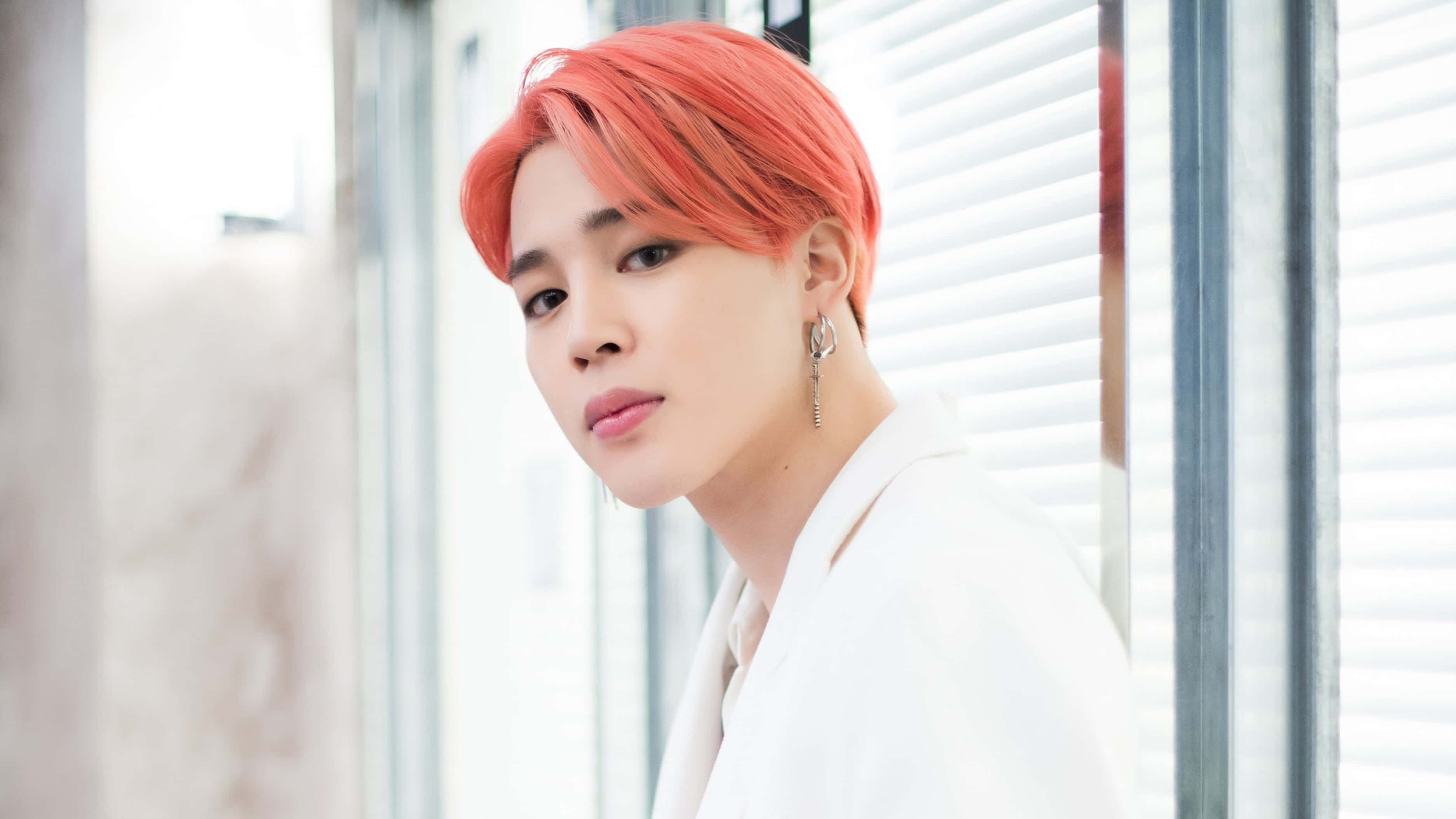 Bts Jimin 桌布 壁紙 — 一個擁有充滿活力的珊瑚色頭髮和銀色耳環的人，在明亮的窗簾旁穿著白色西裝外套，溫柔地凝視著。