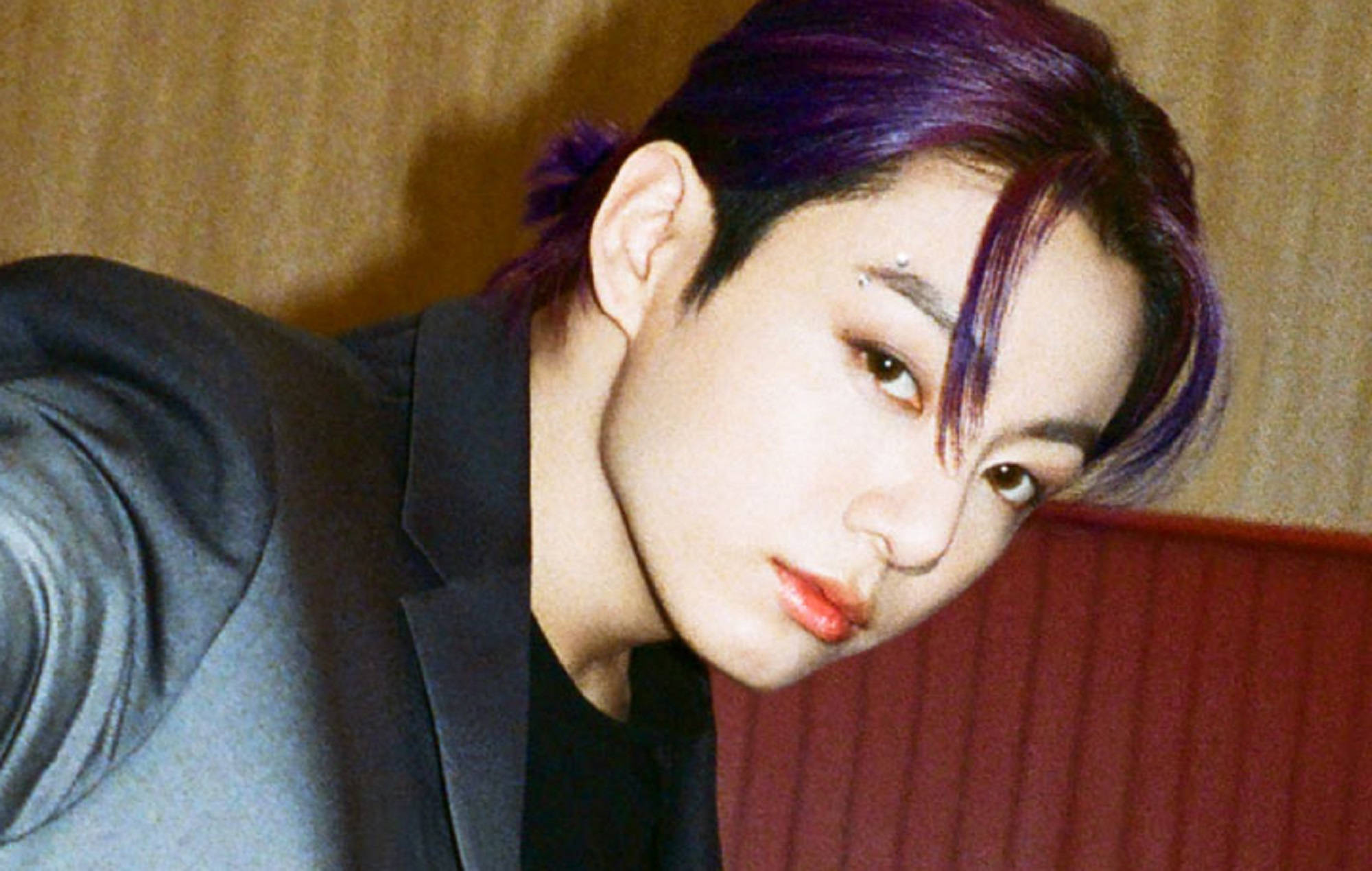 Jungkook 桌布 壁紙 — 一個擁有充滿活力的紫色頭髮和紅唇的人，穿著深色西裝，在溫暖的木質和紅色背景下凝視著。