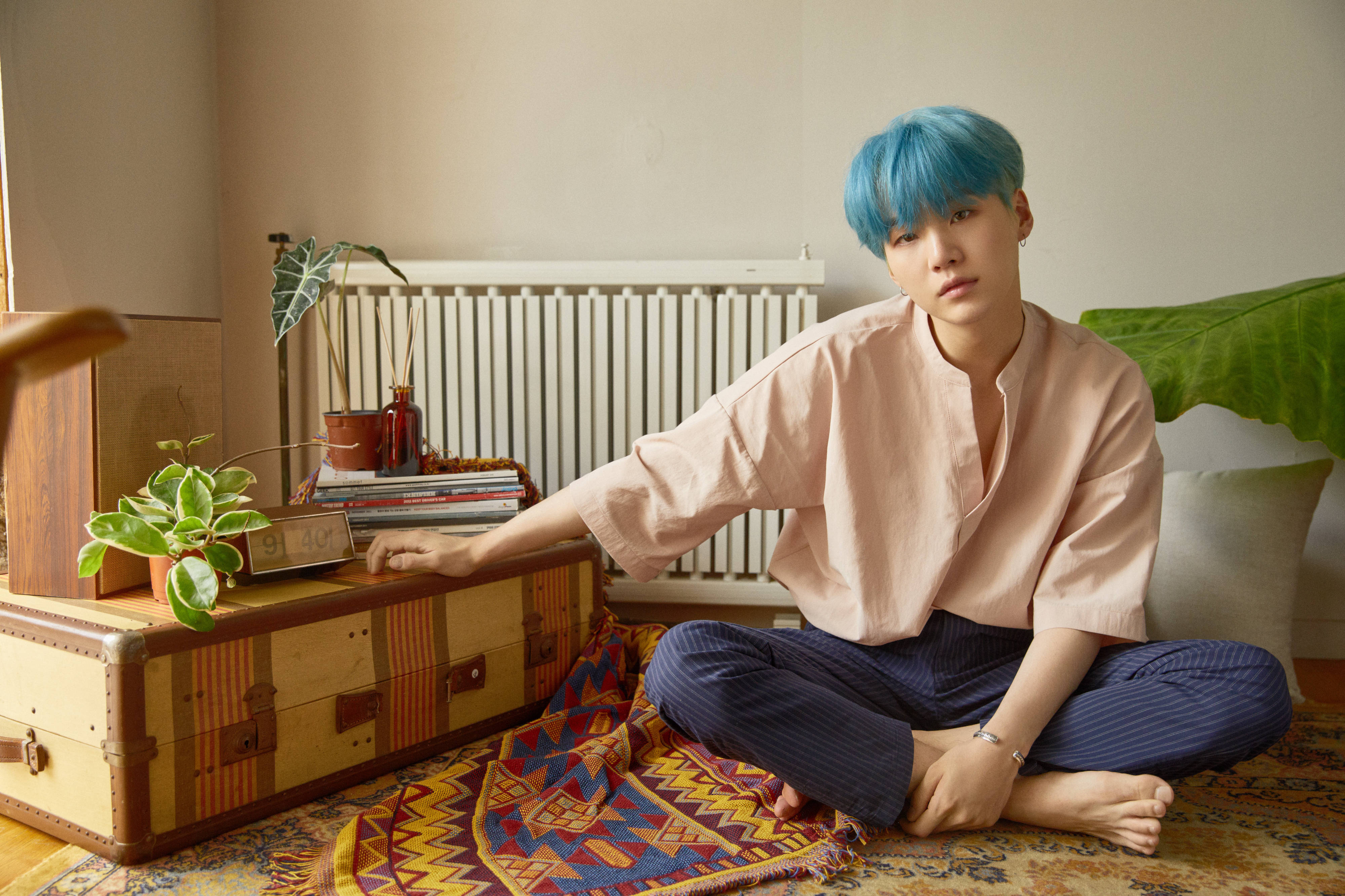 Suga Bts 桌布 壁紙 — 一個擁有充滿活力的藍色頭髮的人，在溫暖照明的房間裡，盤腿坐在有圖案的地毯上，旁邊放著一個復古手提箱。
