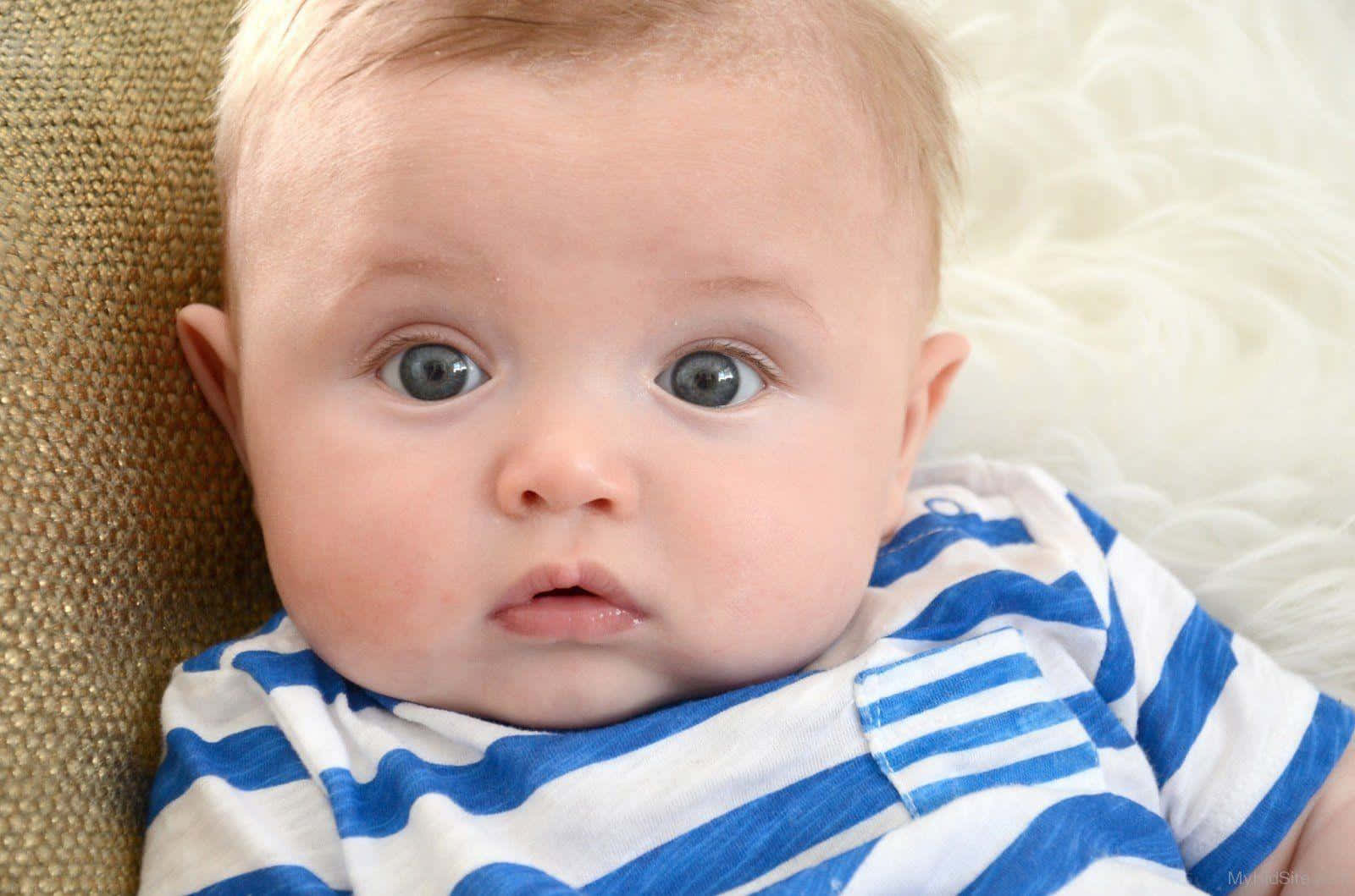 cute baby 桌布 壁紙 — 一個擁有寬闊藍眼睛的嬰兒穿著藍白條紋襯衫，倚靠在有紋理的米色沙發上的特寫。
