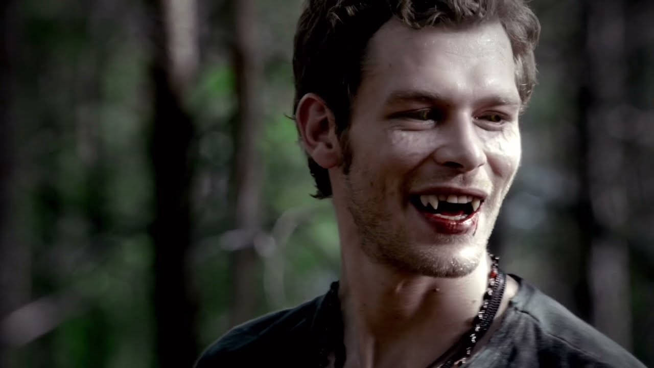 Vampire Diaries 桌布 壁紙 — 一個擁有尖牙和紅唇的吸血鬼在黑暗的森林中猙獰地咧嘴笑。