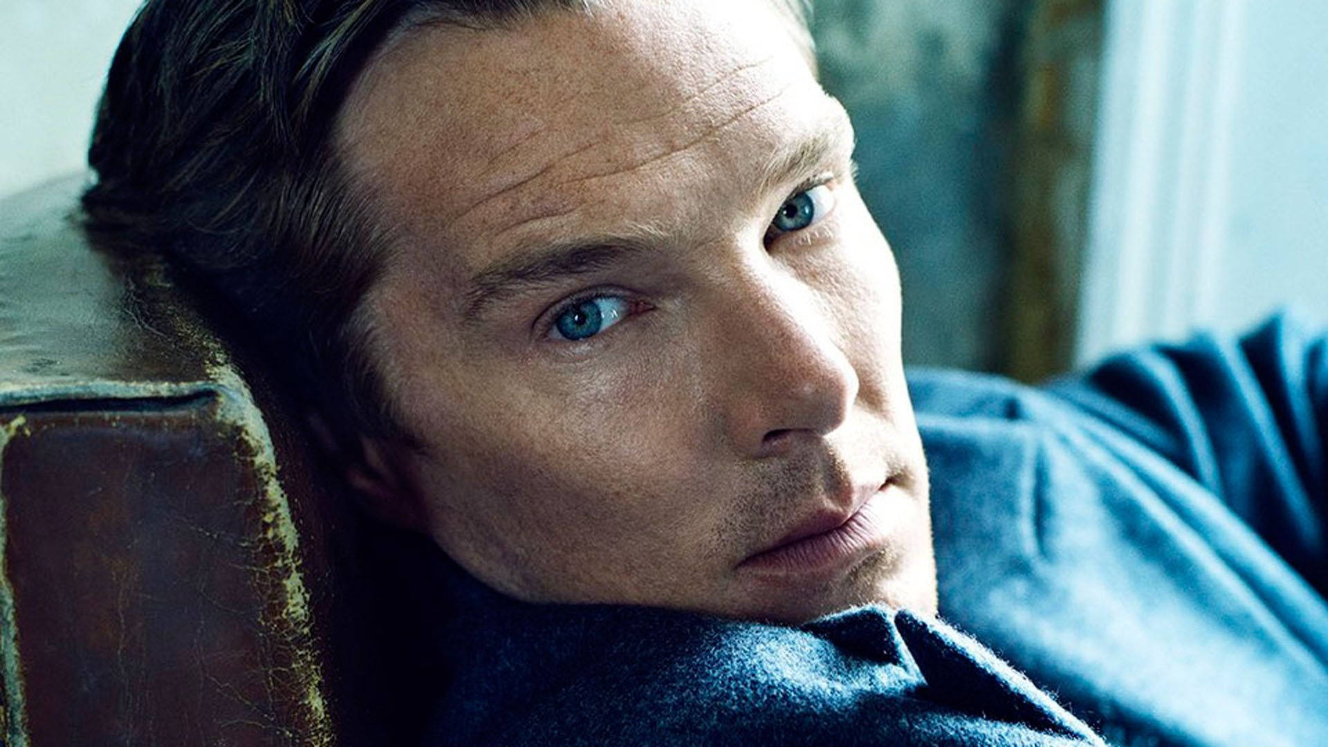 benedict 桌布 壁紙 — 一個擁有引人注目藍眼睛的男人，將頭靠在磨損的皮革扶手上，在柔和光線的室內若有所思地凝視。