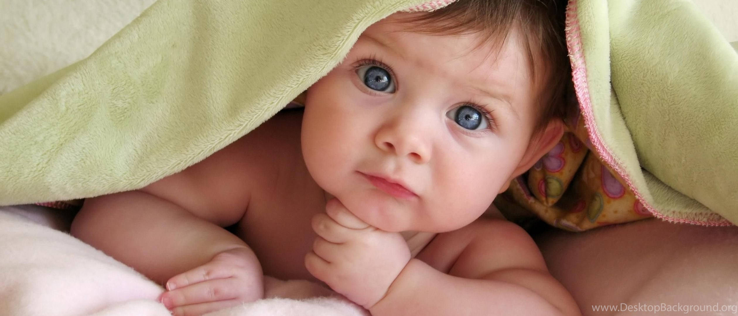 Funny Baby 桌布 壁紙 — 一個擁有引人注目藍色眼睛的嬰兒，從柔軟的綠色毯子下探出頭來，毯子放在蓬鬆的白色表面上。